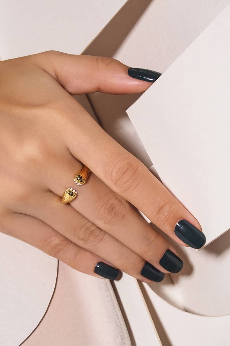 A hand with dark nails adorned with a gold ring featuring cat's paws design, resting on textured paper.