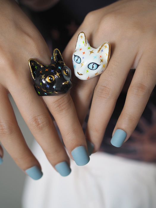 Two ring-shaped cats' heads on each hand. The black cat ring has yellow eyes and a face full of colourful, twinkle-shaped details, while the white cat ring has blue eyes and a face full of colourful, twinkle-shaped details.