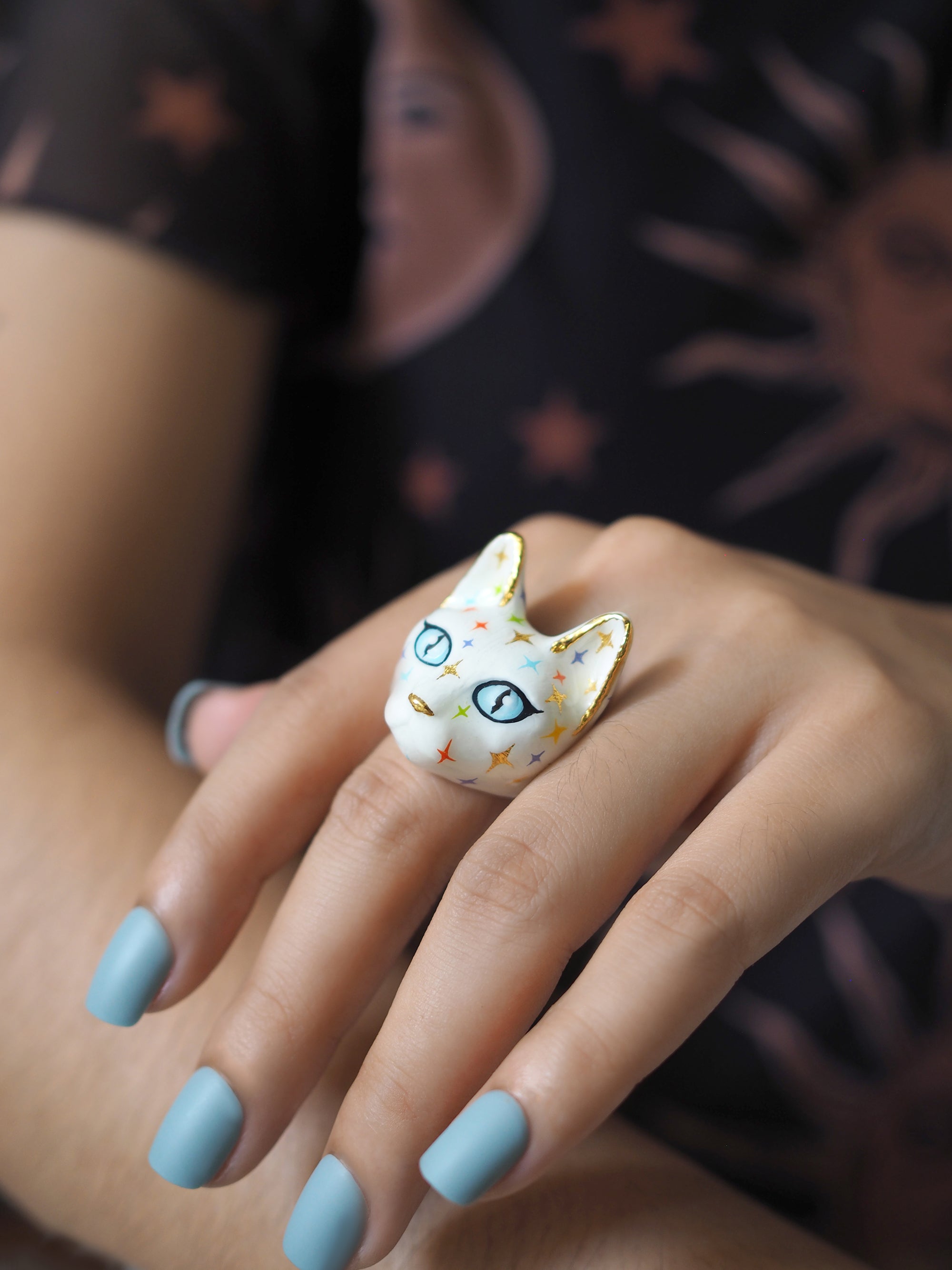 A hand wears a white cat ring with blue eyes and a face full of colourful twinkle-shaped details.