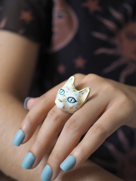 A hand wears a white cat ring with blue eyes and a face full of colourful twinkle-shaped details.