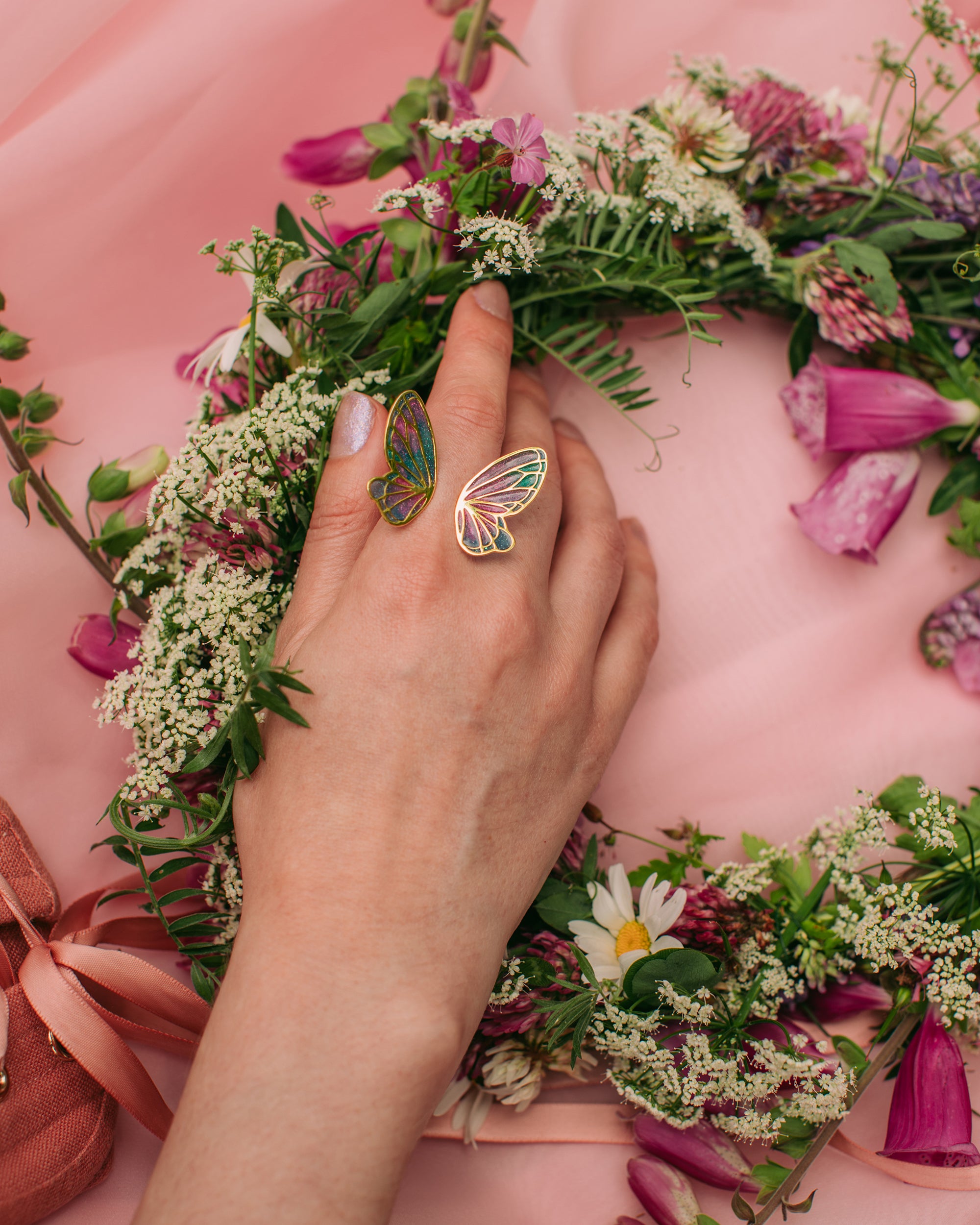 Close-up of a colorful butterfly wing ring on a finger, with pink blossoms in the background.