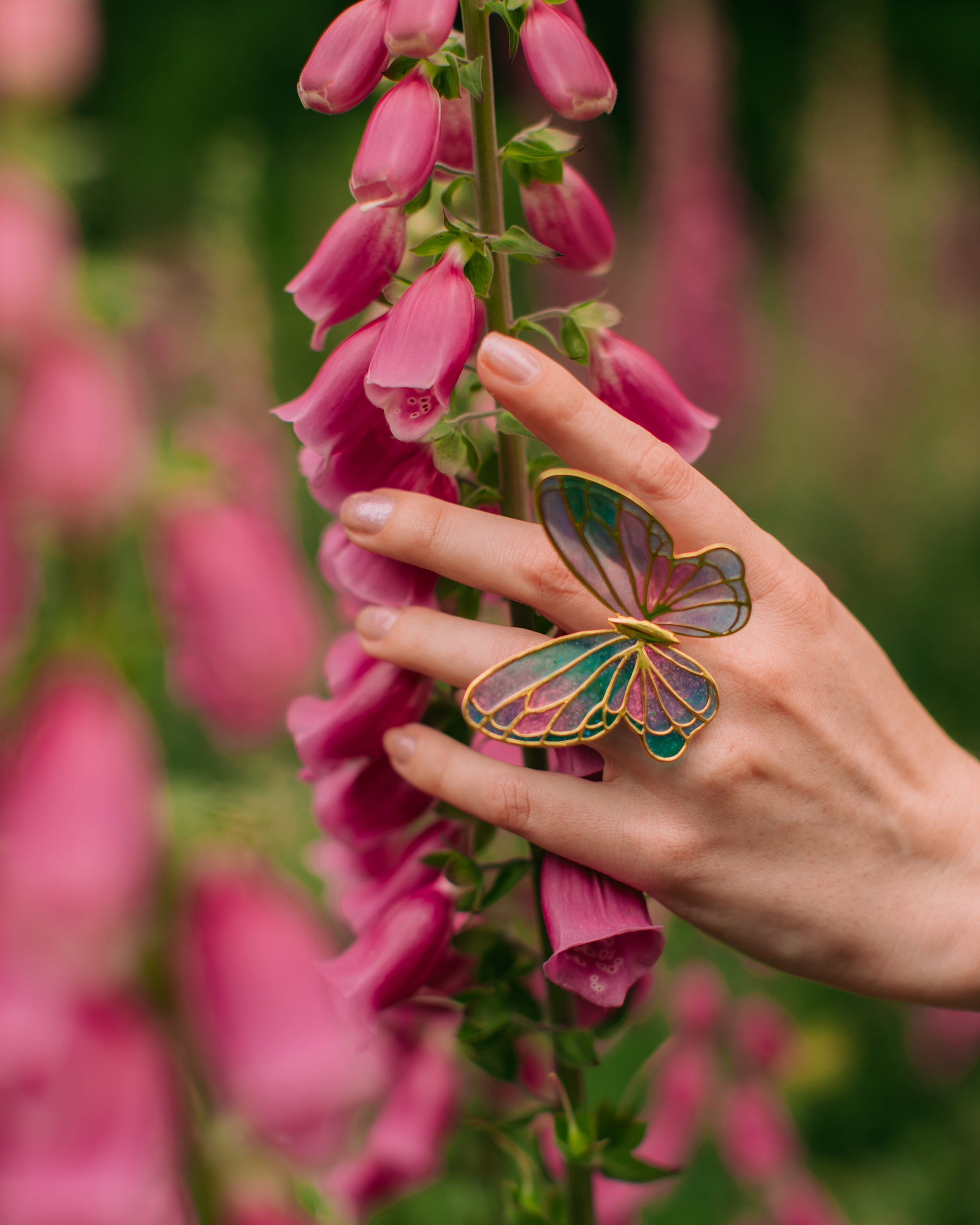 Close-up of a colorful butterfly ring on a finger, with pink blossoms in the background.