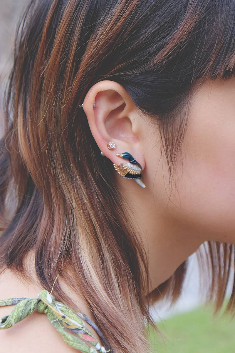 A close-up of a woman's ear featuring a mallard duck earring, with short hair.