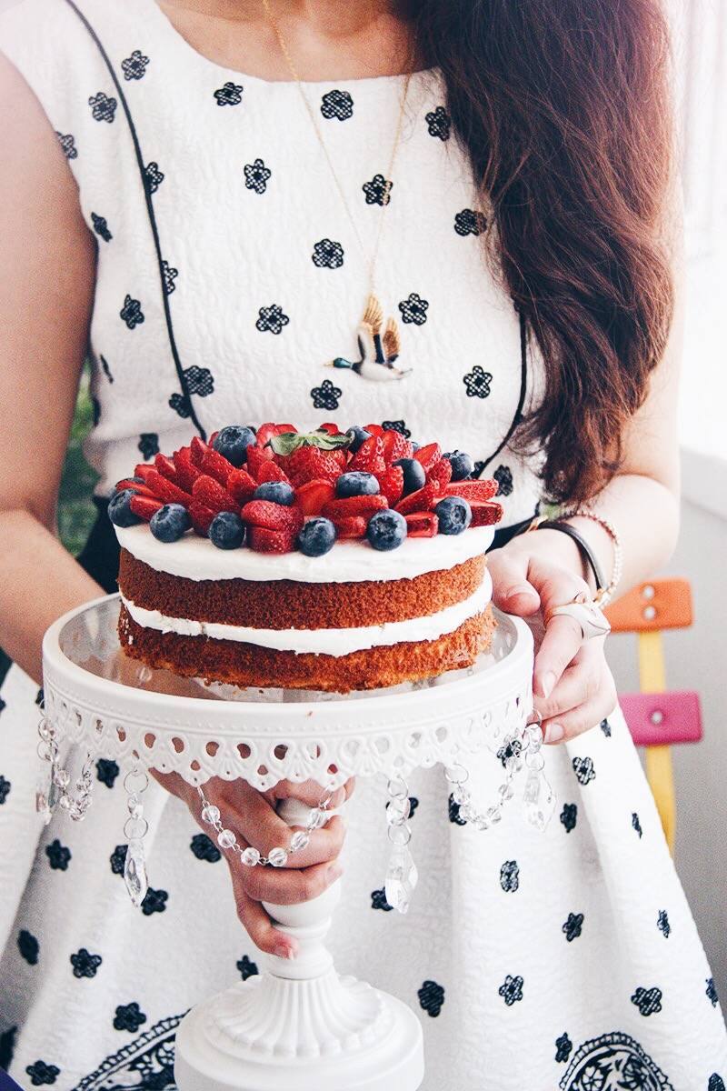 A woman in a patterned white dress holds a layered cake adorned with strawberries and blueberries, wearing a mallard necklace.