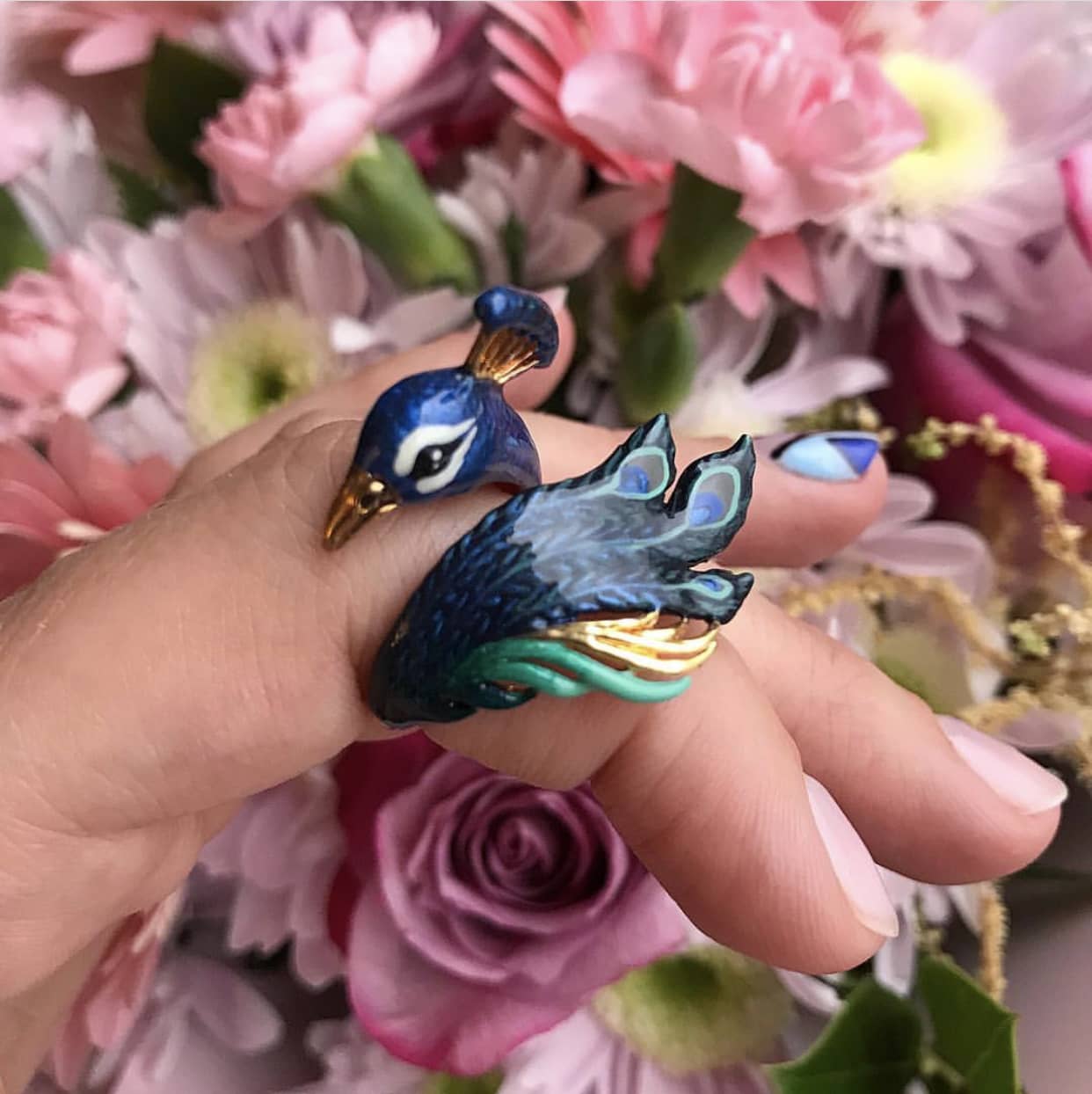 Close up of a hand wearing a peacock ring with intricate details surrounded by vibrant flowers.