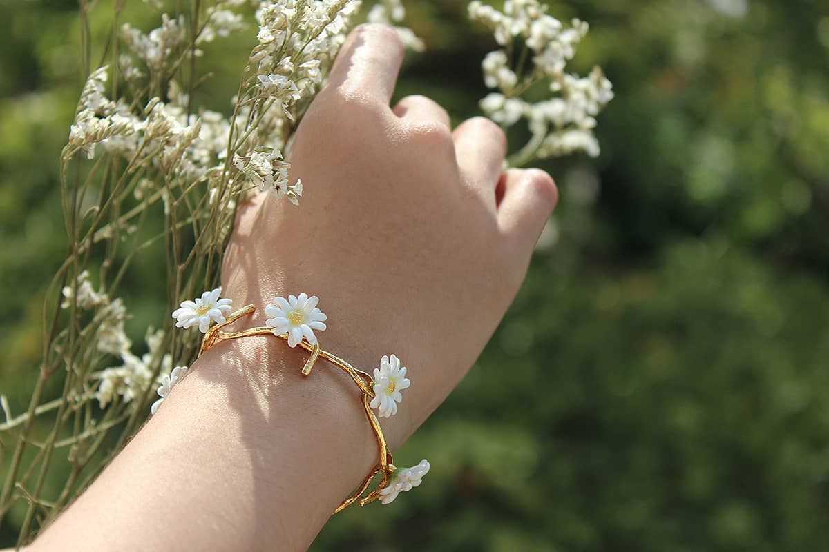 Close-up of a hand showcasing a beautiful gold bracelet with white daisies, surrounded by flowers.