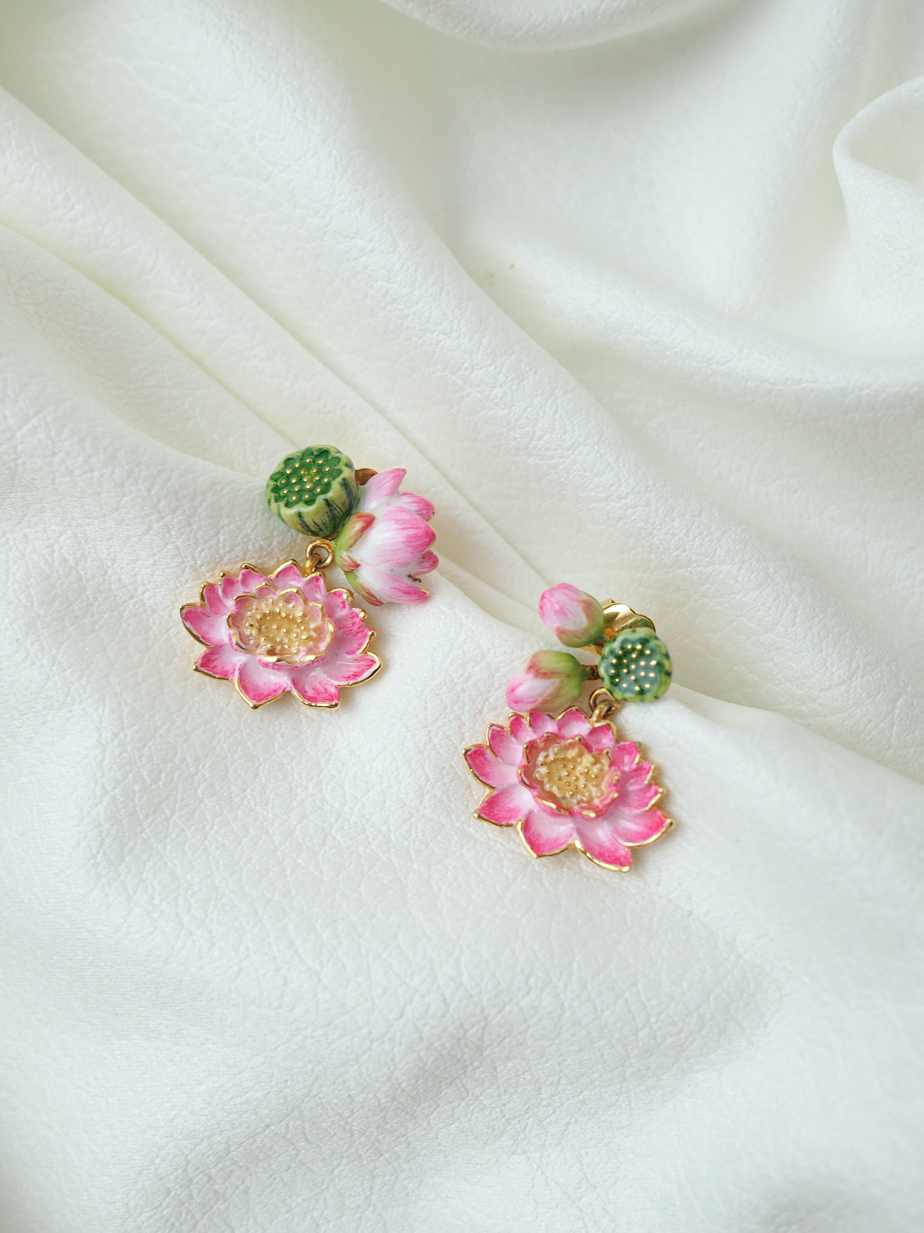 Beautiful floral earrings featuring pink lotus flowers with green decorative elements placed on a white fabric.