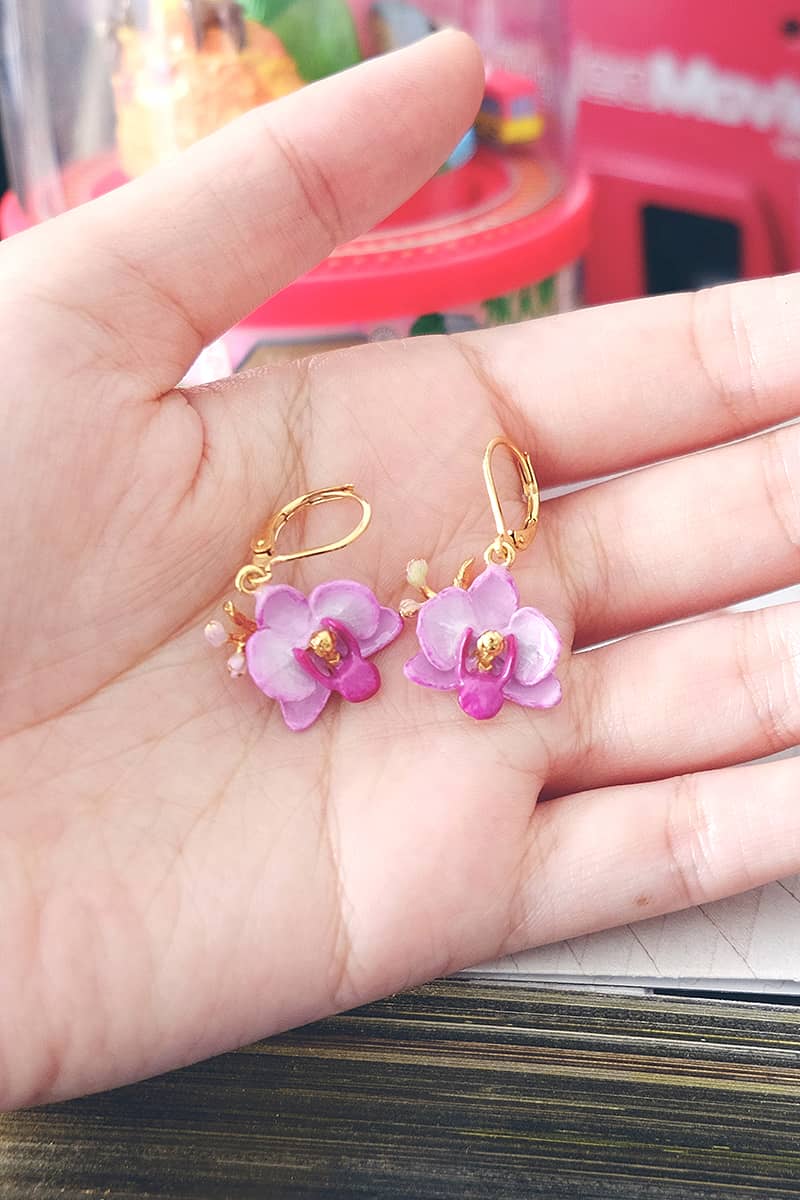 Close up of floral earrings featuring purple phalaen design held in a person's hand.