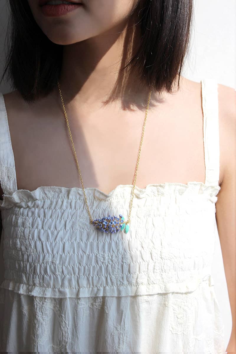 A woman wears a floral necklace, featuring a cluster of lilac flowers and green leaves, on her blouse.