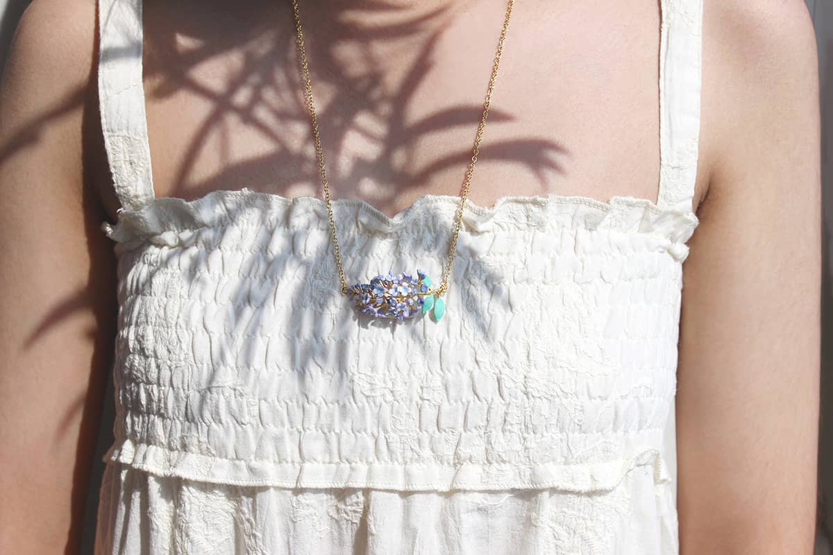 A woman wears a floral necklace, featuring a cluster of lilac flowers and green leaves, ideal for summer fashion.