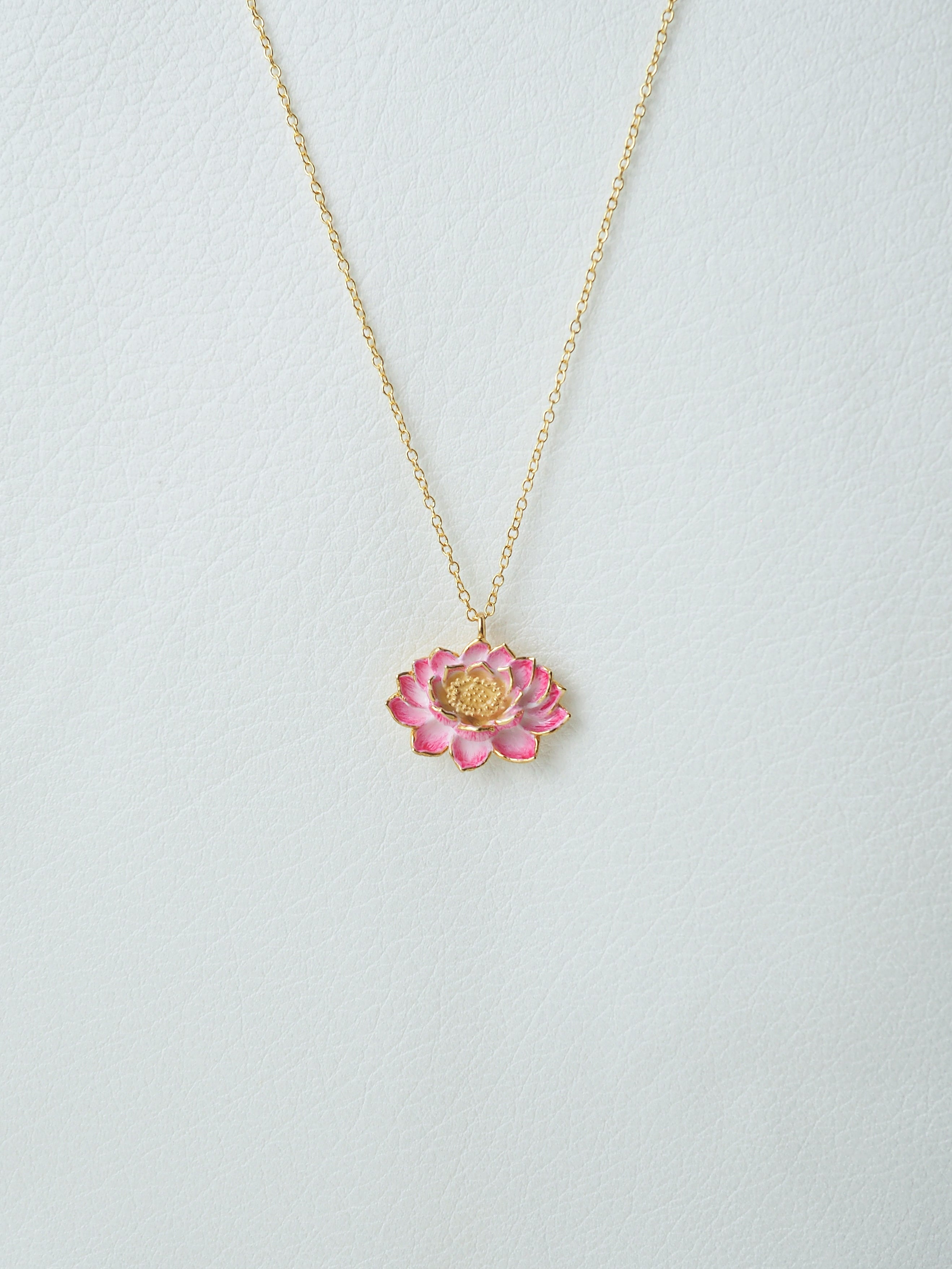 A gold chain necklace features a pink lotus flower pendant, intricately detailed with gold accents in a white background.