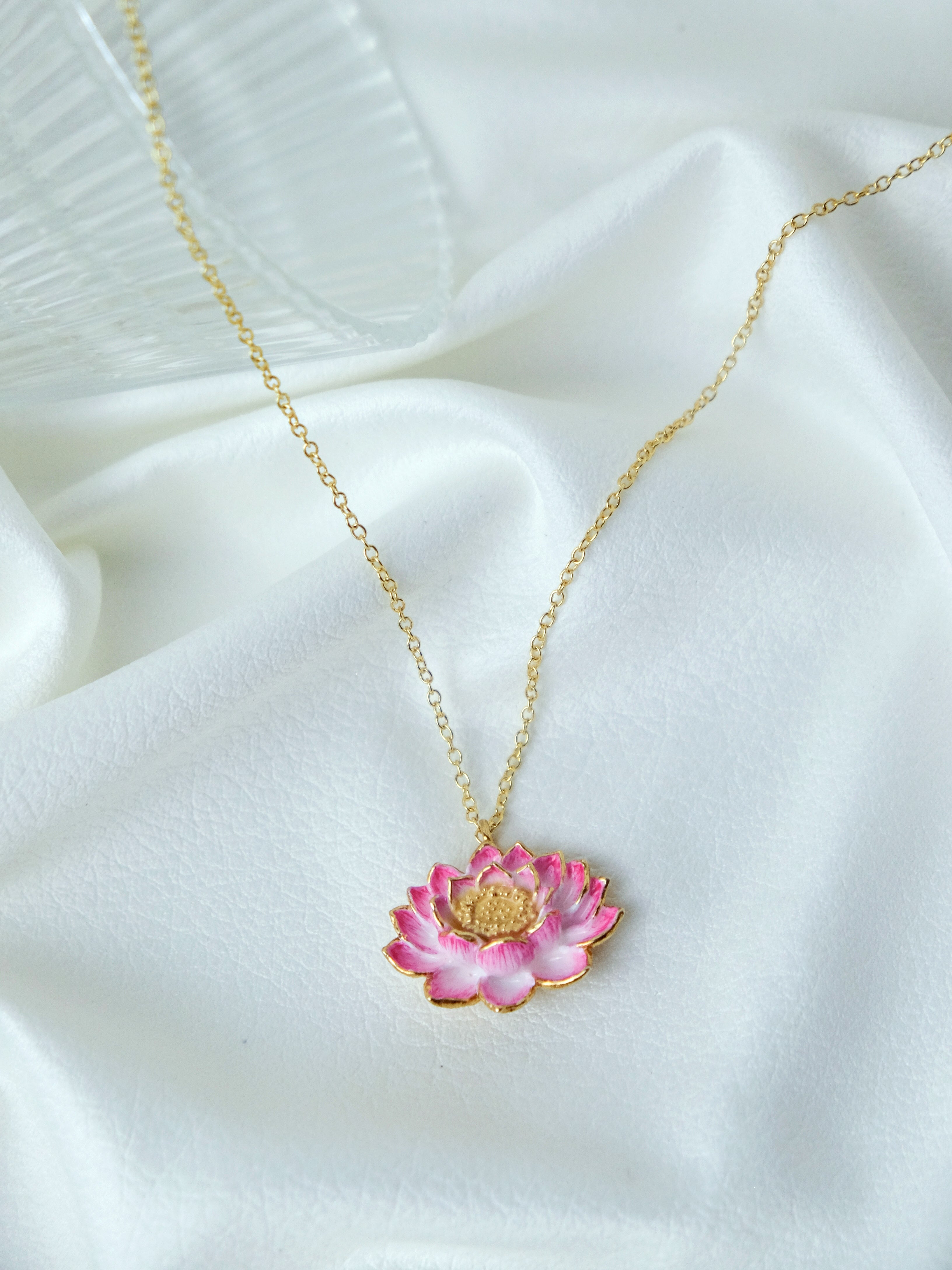 A gold chain necklace features a pink lotus flower pendant, intricately detailed with gold accents in a white fabric.