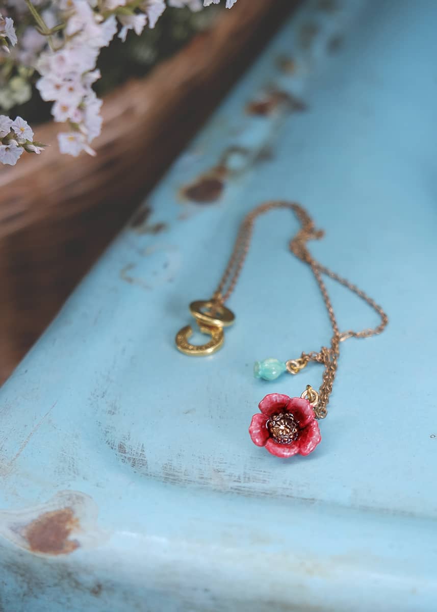 Gold necklace featuring a red poppy pendant and a small green accent, placed on blue background.