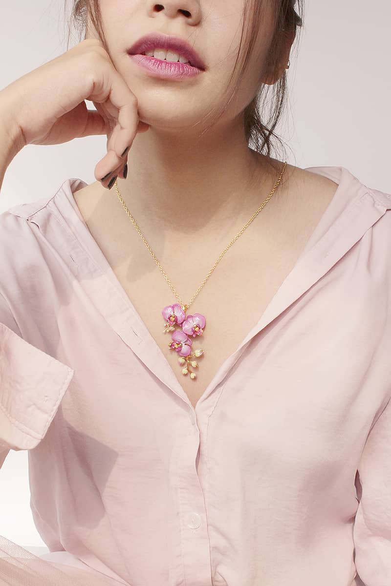 Woman wearing a gold necklace with purple flower details and a light pink shirt.