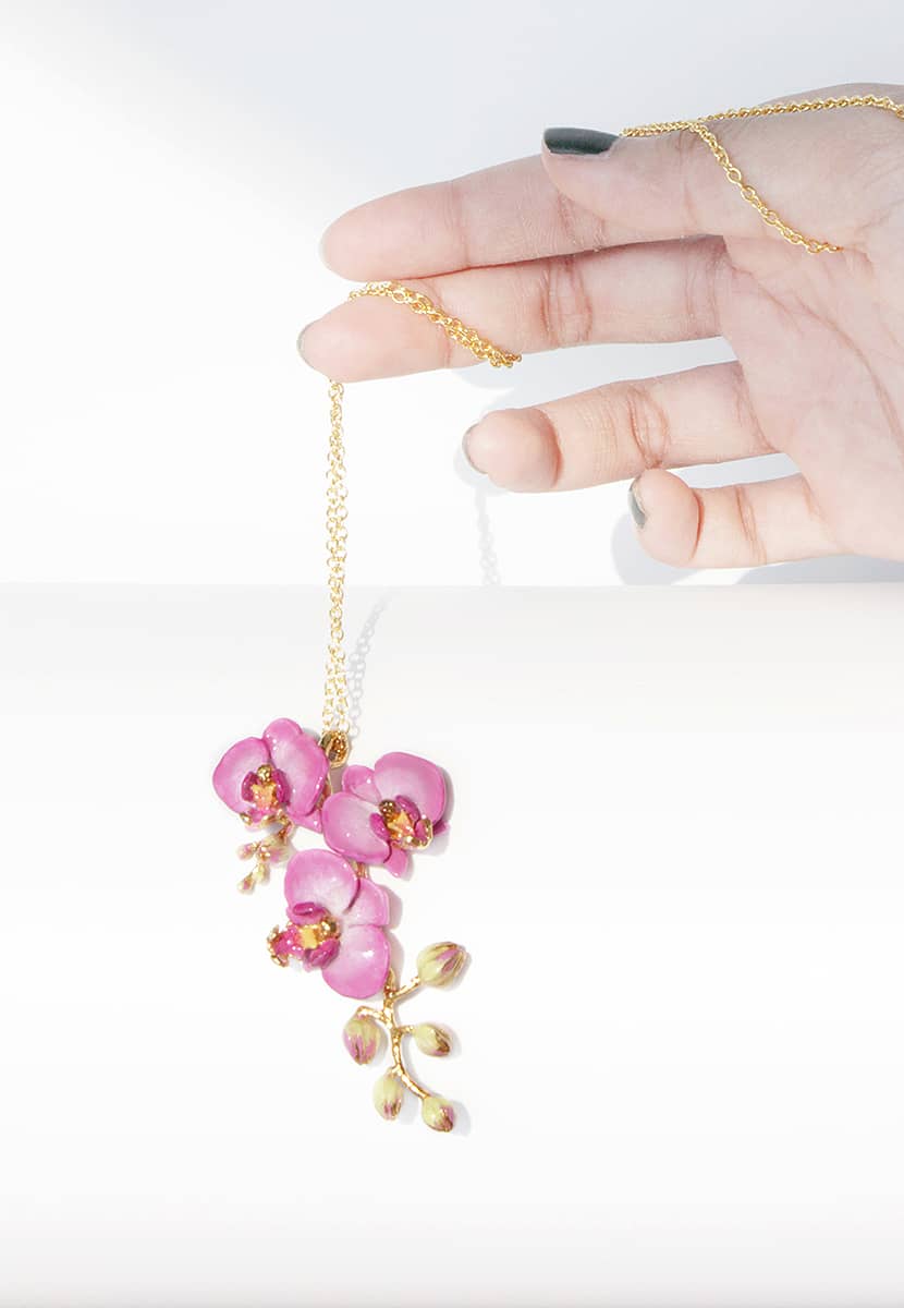 Close up of a hand holding a gold necklace with three purple flowers and golden accents pendant.