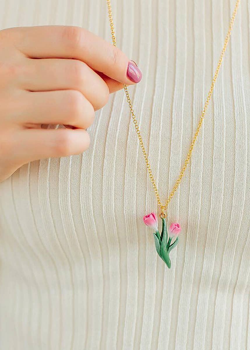 Hand holding a gold necklace with pink tulip pendant against a ribbed, light-colored top.