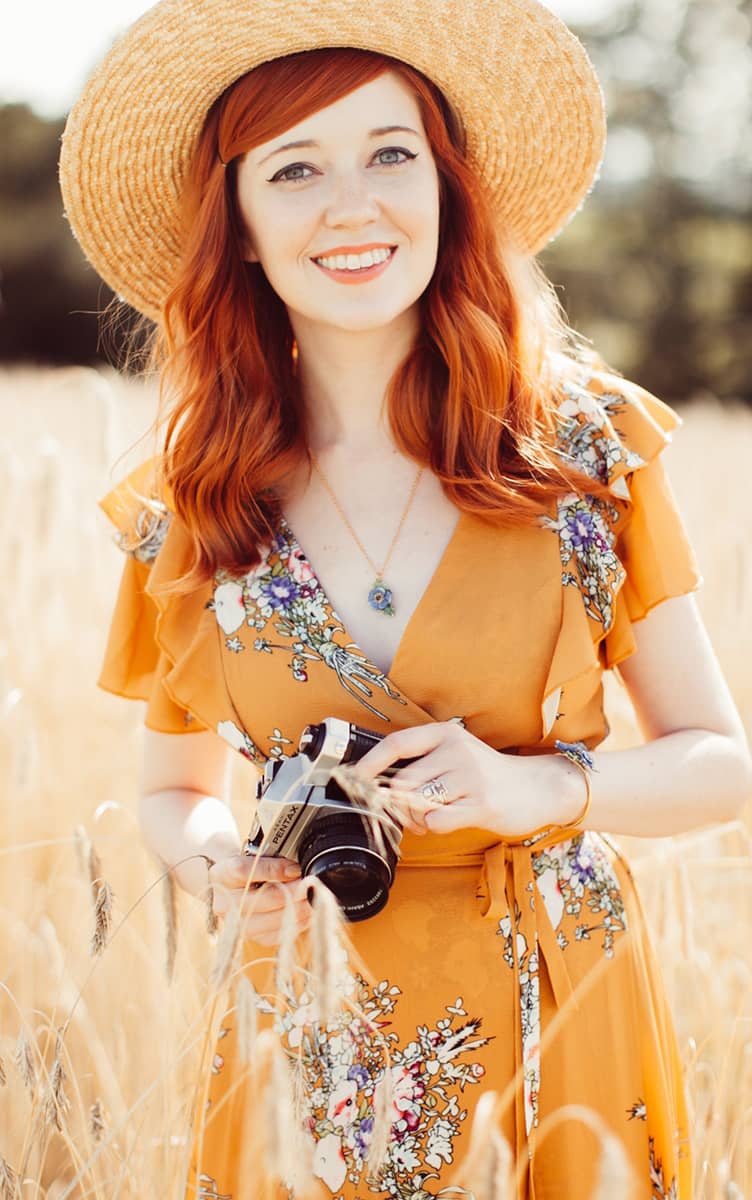 A woman with red hair in a floral dress wearing a stylish flower necklace standing in a sunny field.