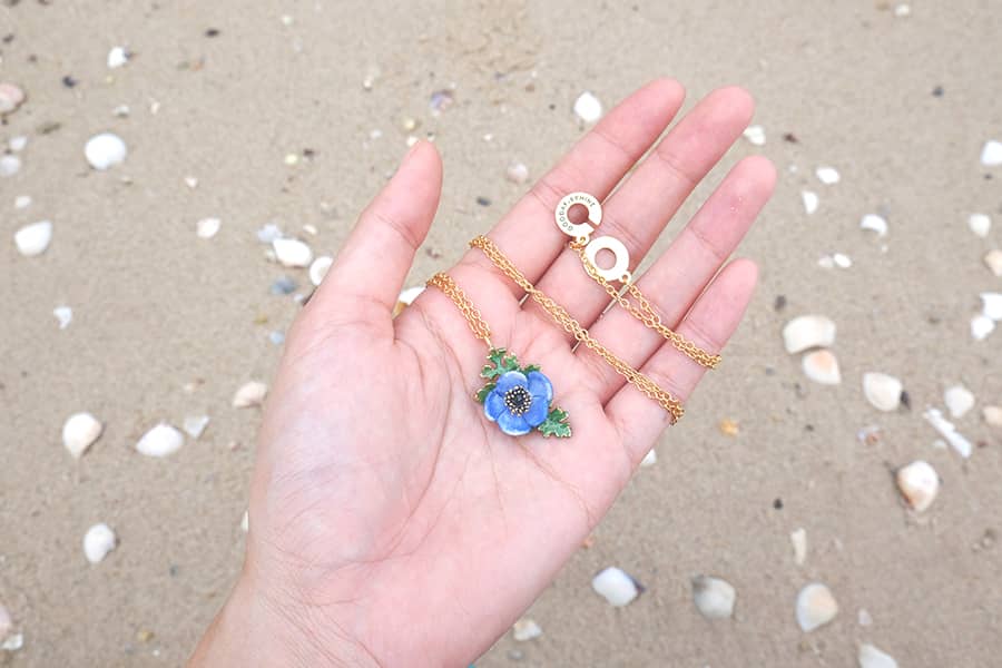 Close-up of a hand holding necklace with a purple flower and green  leaves on sandy beach.