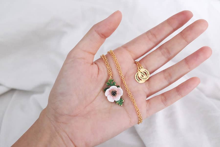 Close-up of a hand holding a stunning necklace featuring a light pink, white flower and green leaves pendant.