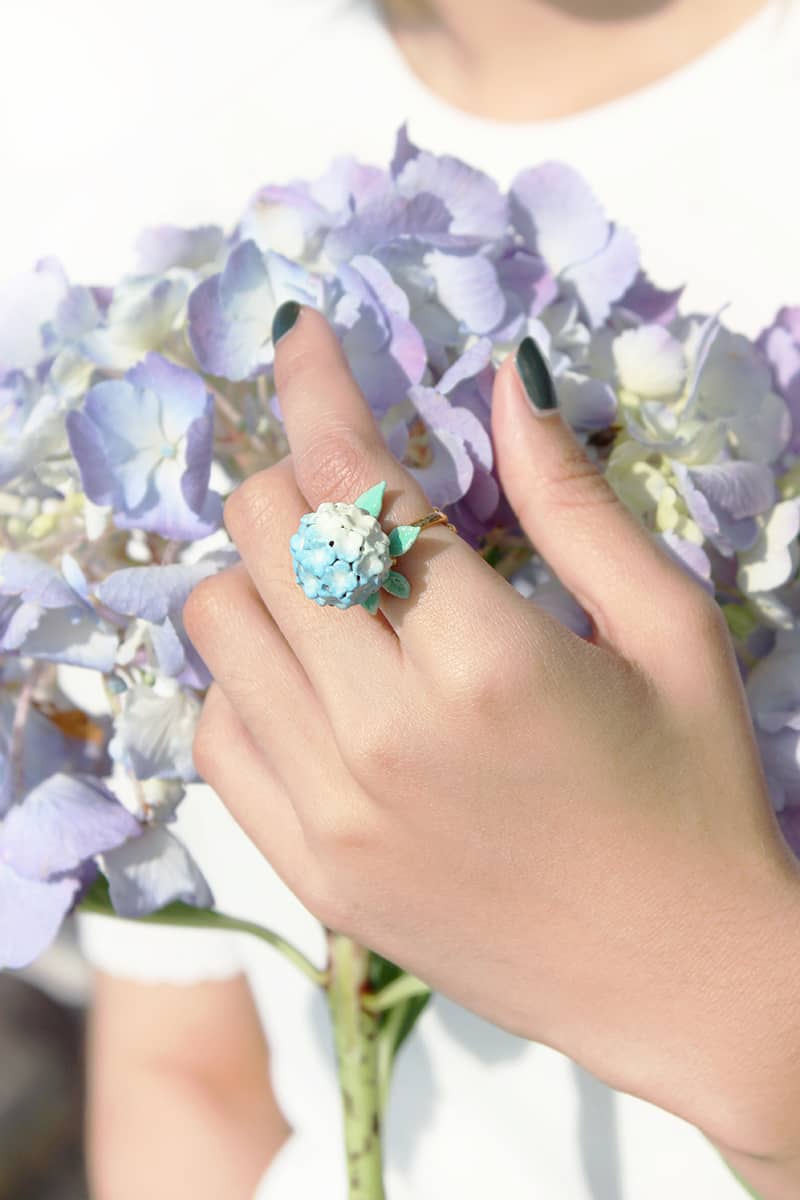 Close-up of a hand wearing a floral ring with blue and white flowers, holding a bouquet of hydrangeas.