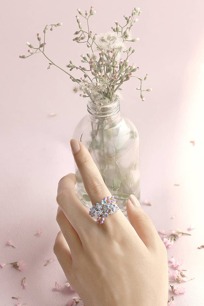 A delicate purple flower ring worn on a hand, with a glass vase of flowers in the background.