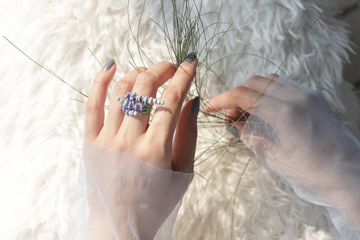 A close-up of a hand with lavender ring, surrounded by green grass, set against a soft white fur background.