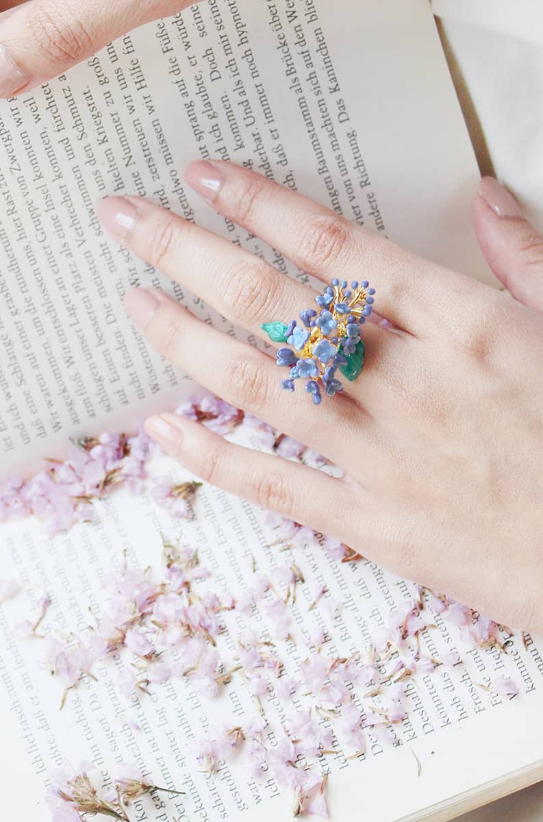 A hand adorned with a purple floral ring rests on an open book, surrounded by delicate pink petals.