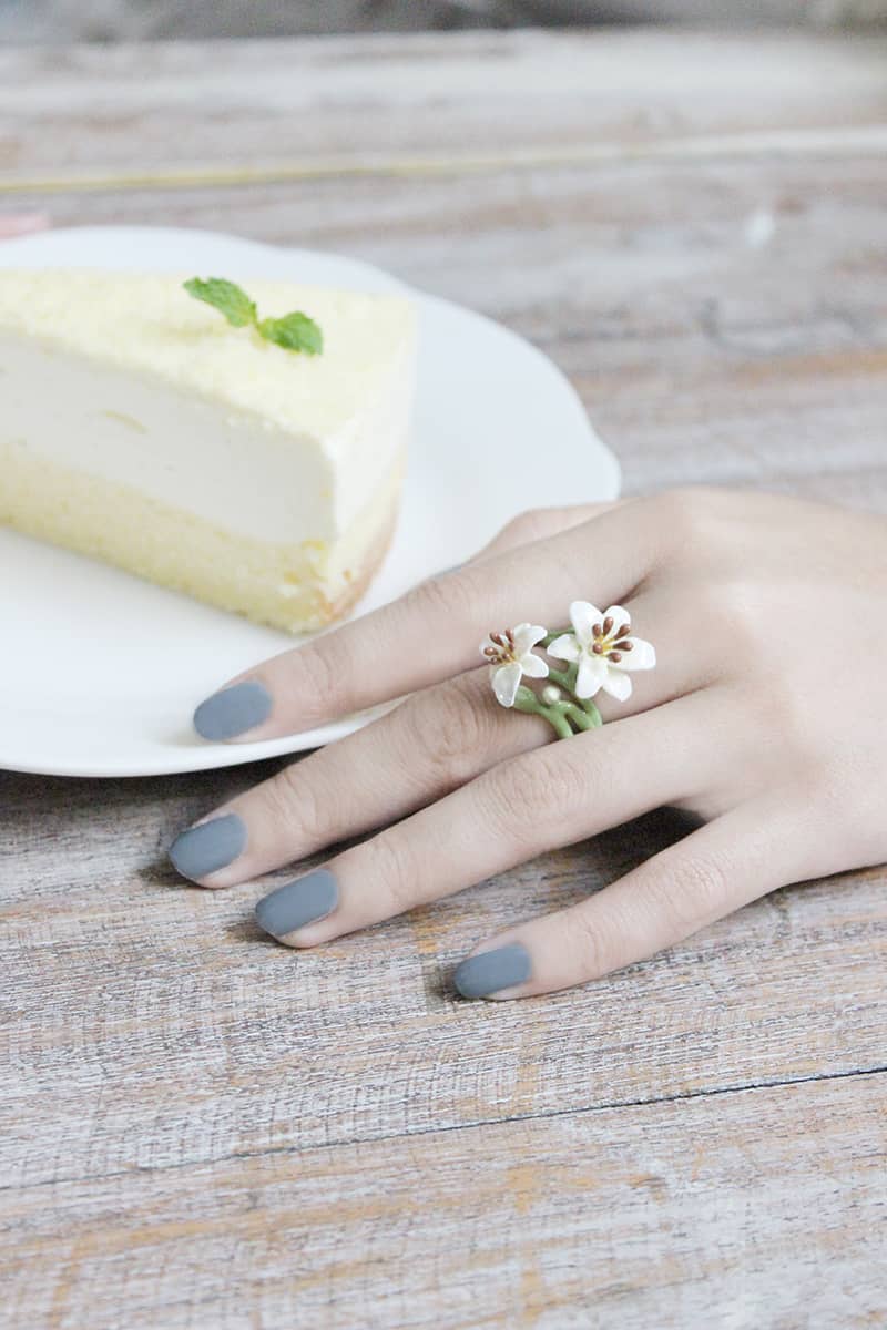 A hand with gray nails adorned with a lily flowers ring rests beside a slice of creamy yellow cake on a plate.