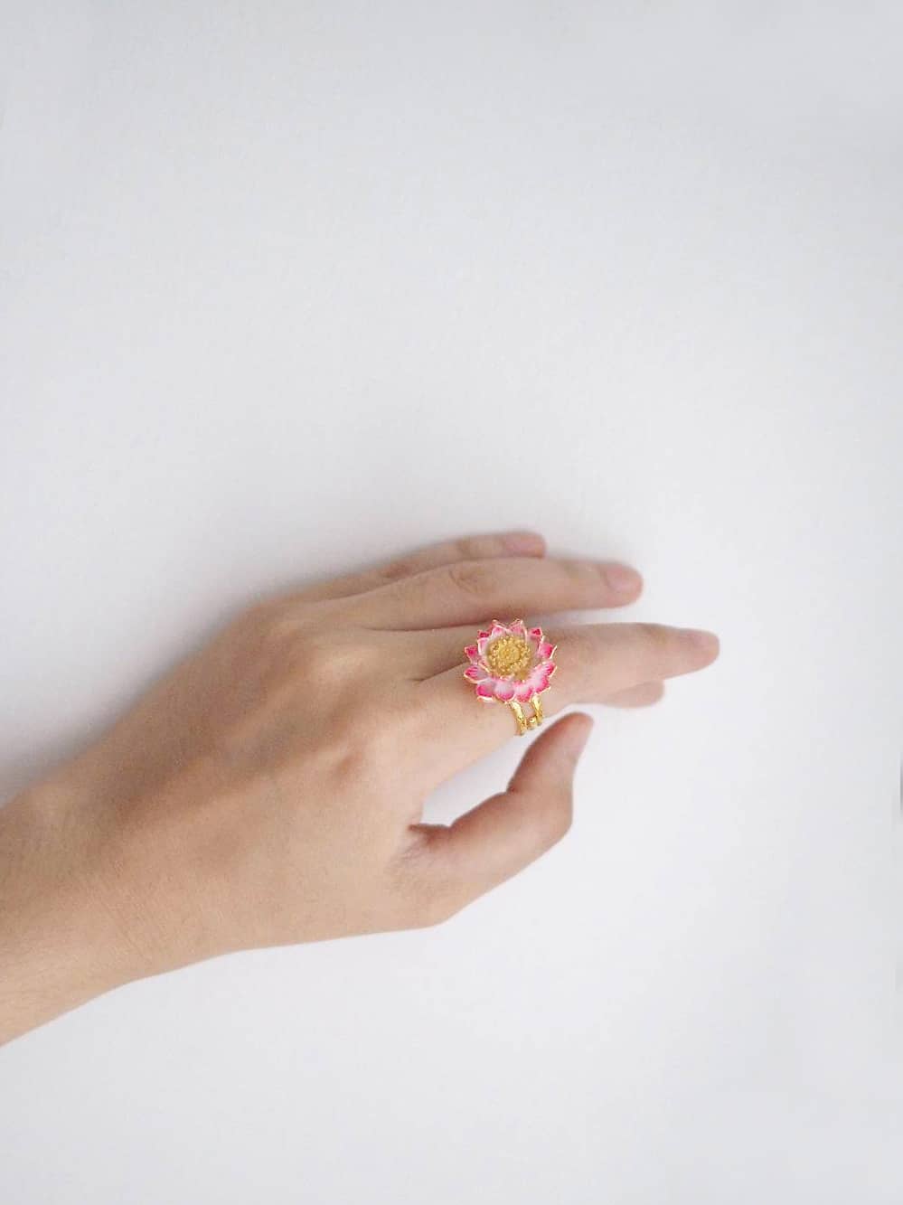 A hand wearing a ring featuring a pink lotus flower design, set against a clean, white background.