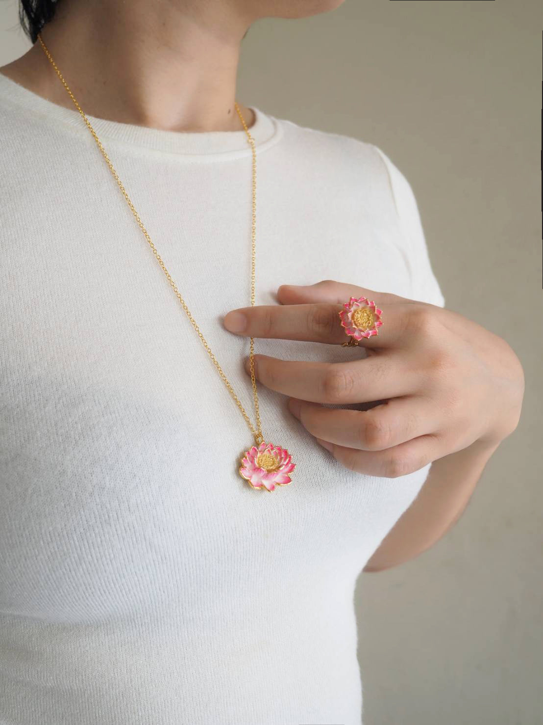 A woman holds a hand to her chest, showing a gold necklace with a pink flower pendant and a matching ring.