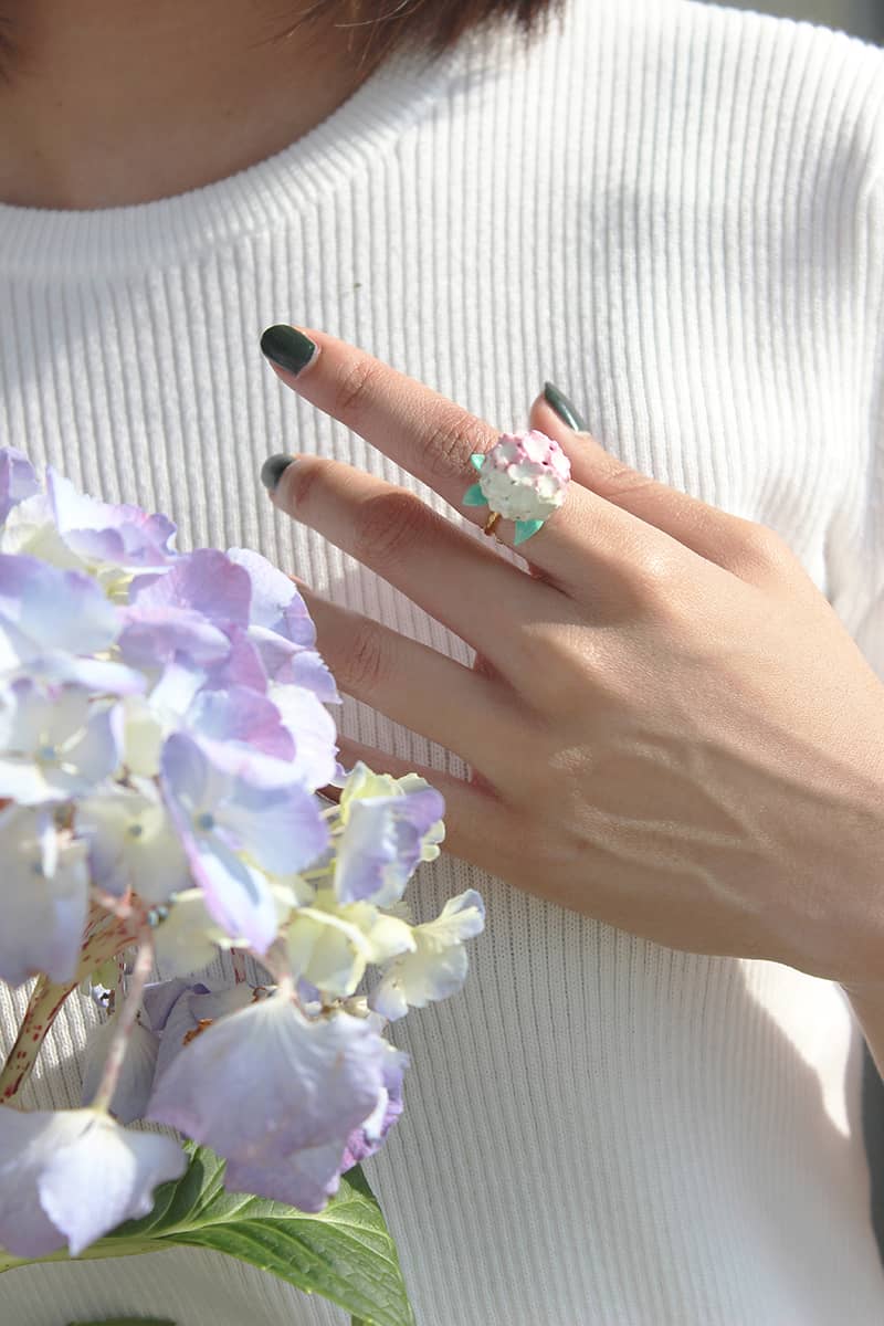 A hand wearing a hydrangea ring next to blooming hydrangeas.