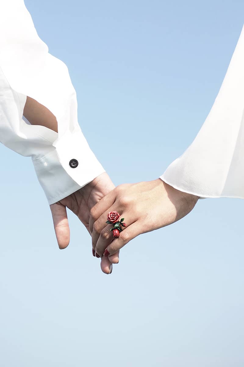 A close-up view of two hands holding each other, one adorned with a rose ring, against a soft blue background.