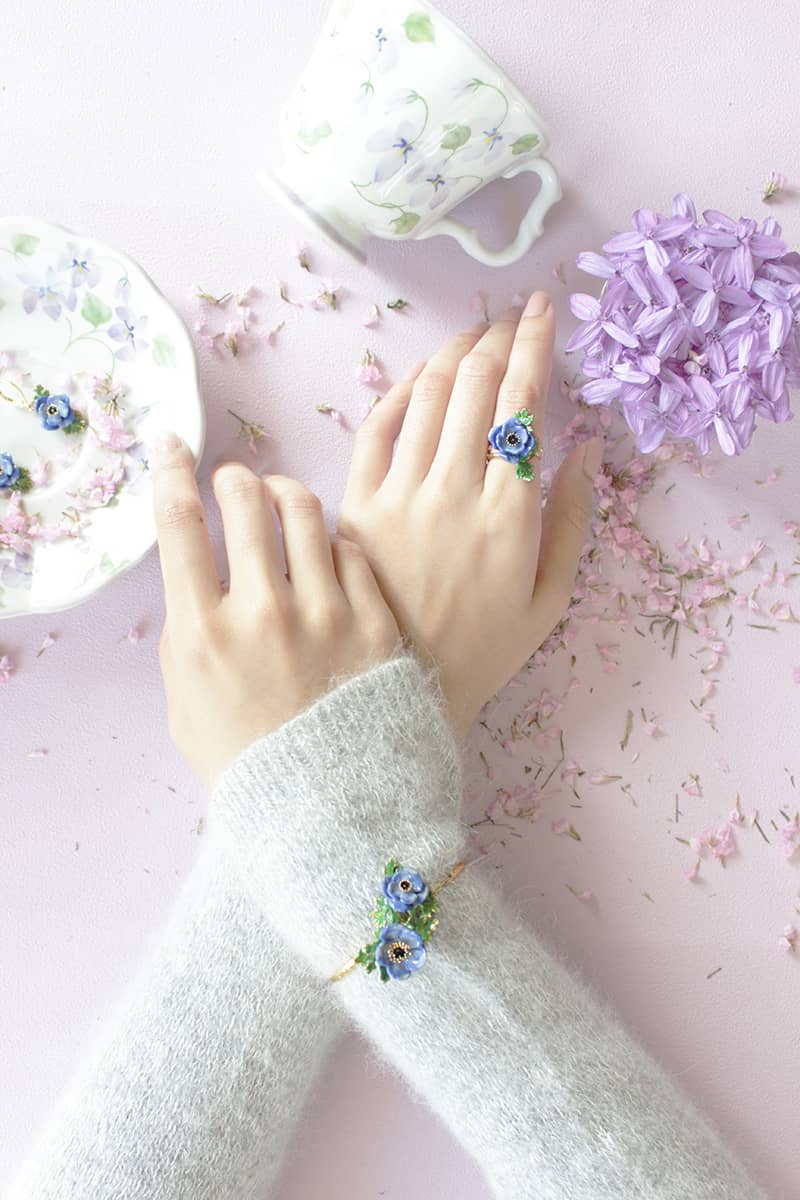 Close-up of a delicate flower ring and flower bangle worn on a hand with a cozy sweater, surrounded by a tea cup and fresh flowers.