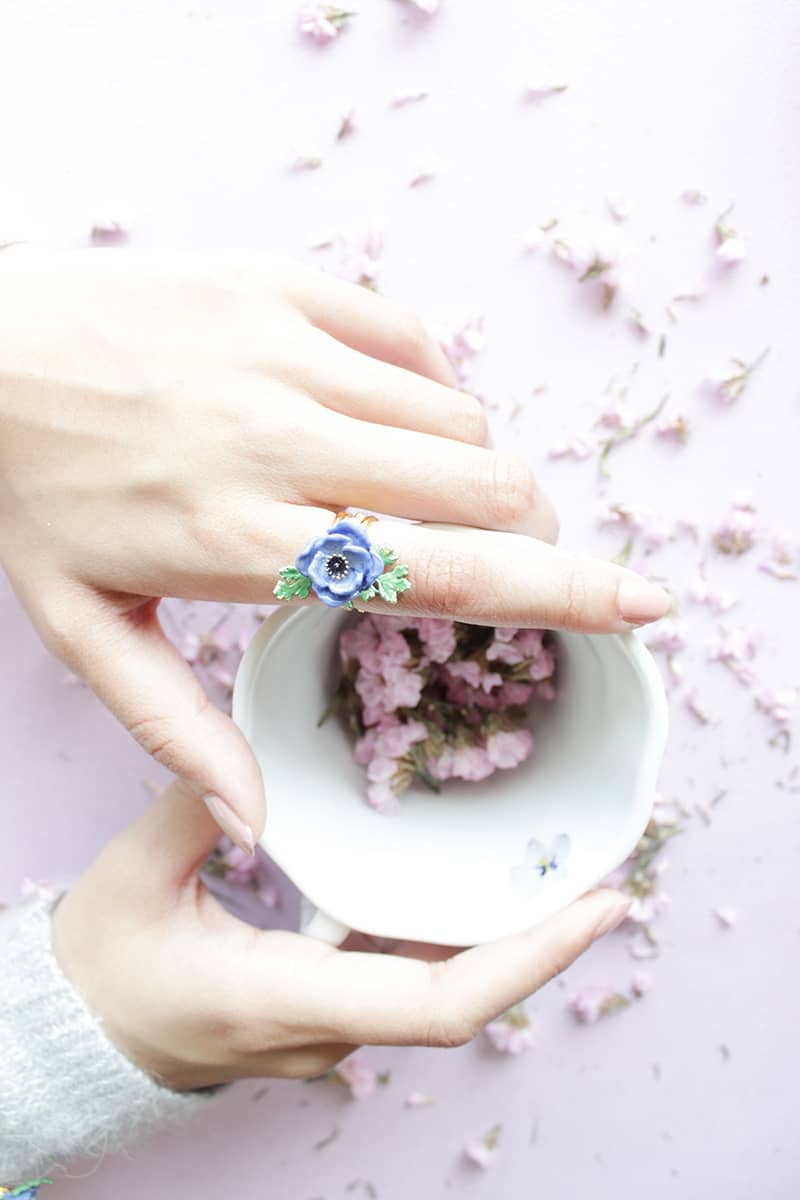 A hand wearing a purple flower ring, holding a white cup filled with pink flowers.