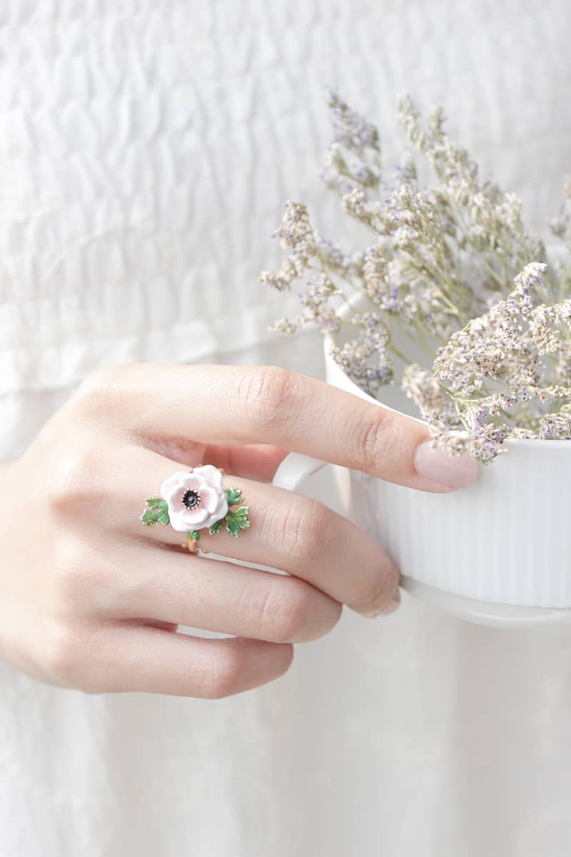 Close-up of a hand adorned with a light pink flower and green leaves ring, gently holding a cup. 