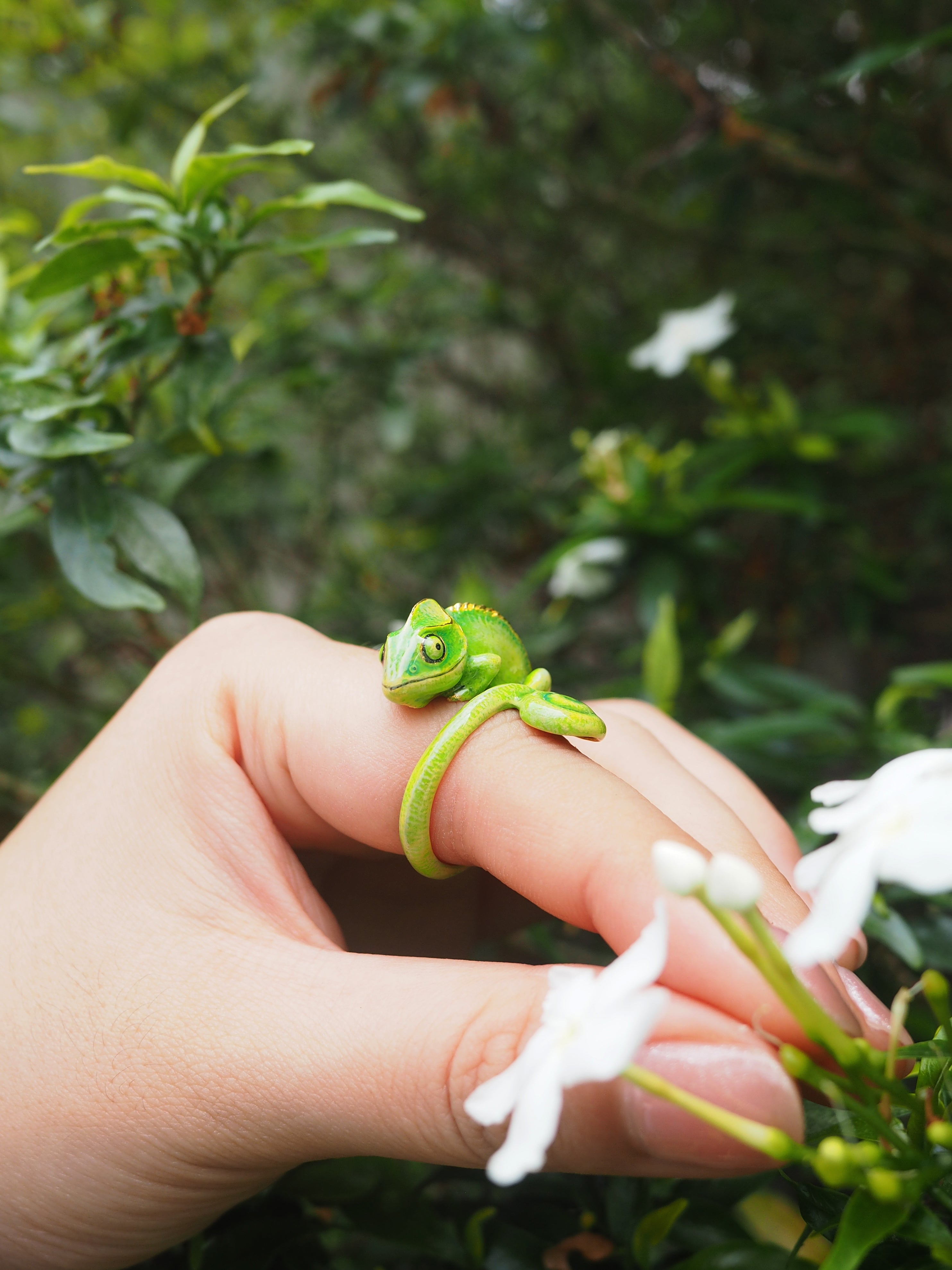 A whimsical chameleon ring on a finger, with vibrant green plants and flowers around.