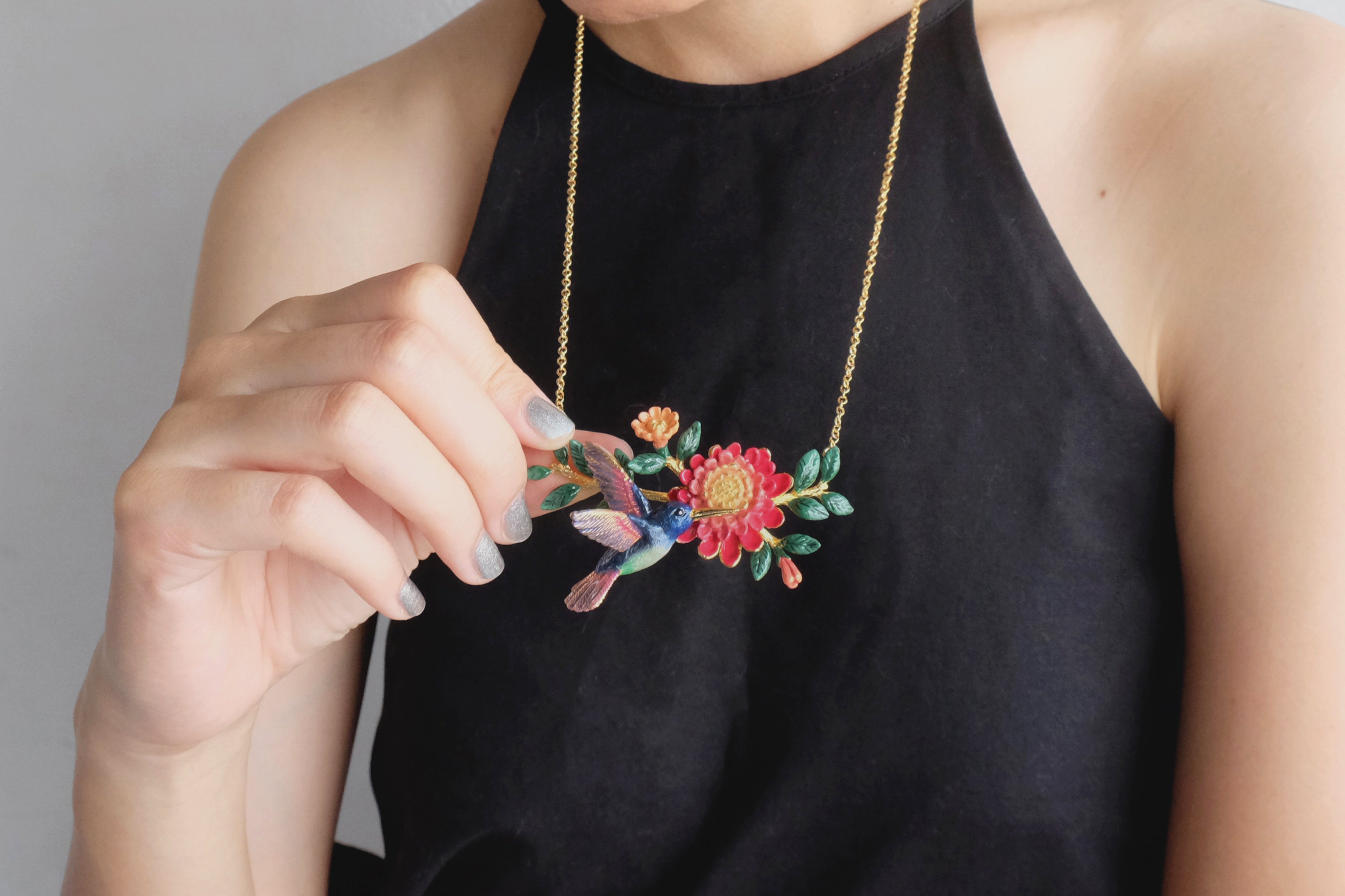 Detailed view of a stunning necklace featuring a hummingbird and colorful flowers, held by a model.