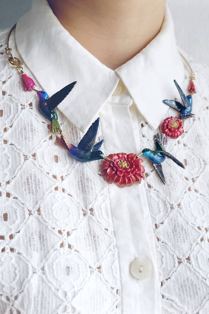 A statement necklace featuring hummingbirds and floral elements, styled with a white lace top.