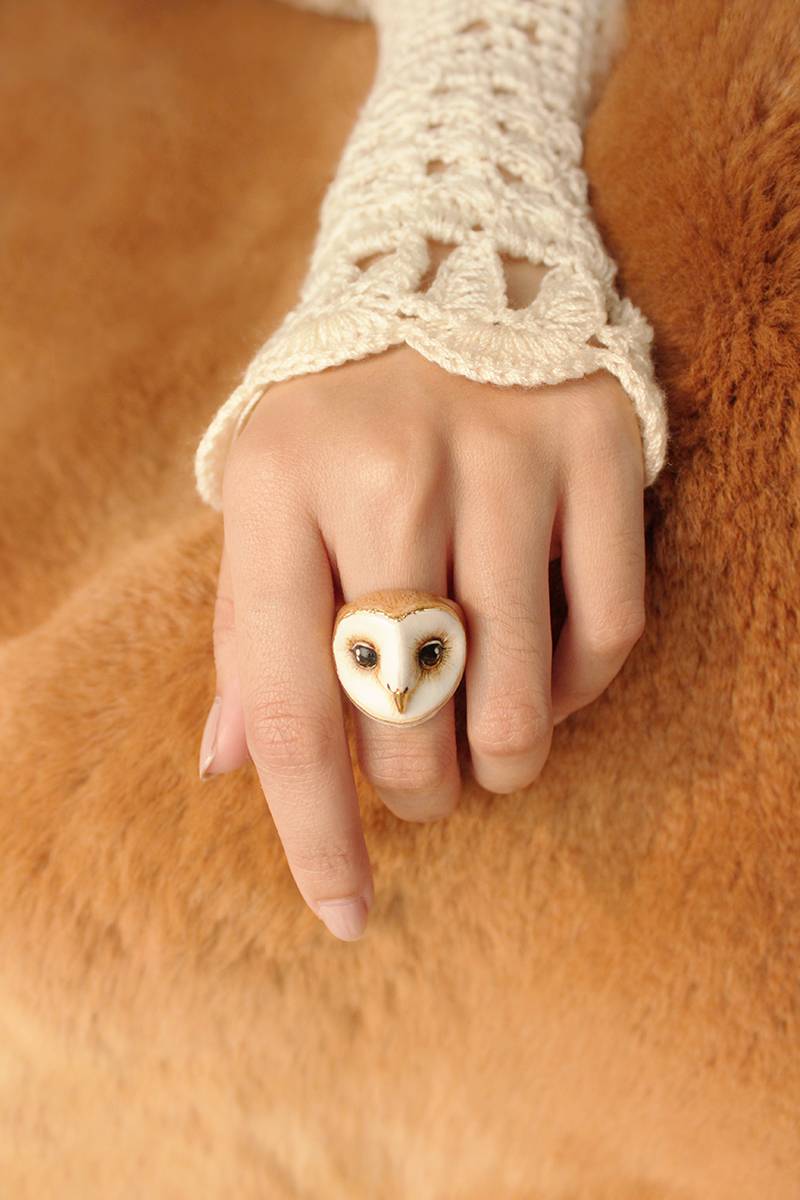 A hand wearing an adorable owl ring, with intricate crochet sleeve, rests on a soft, brown fur surface.