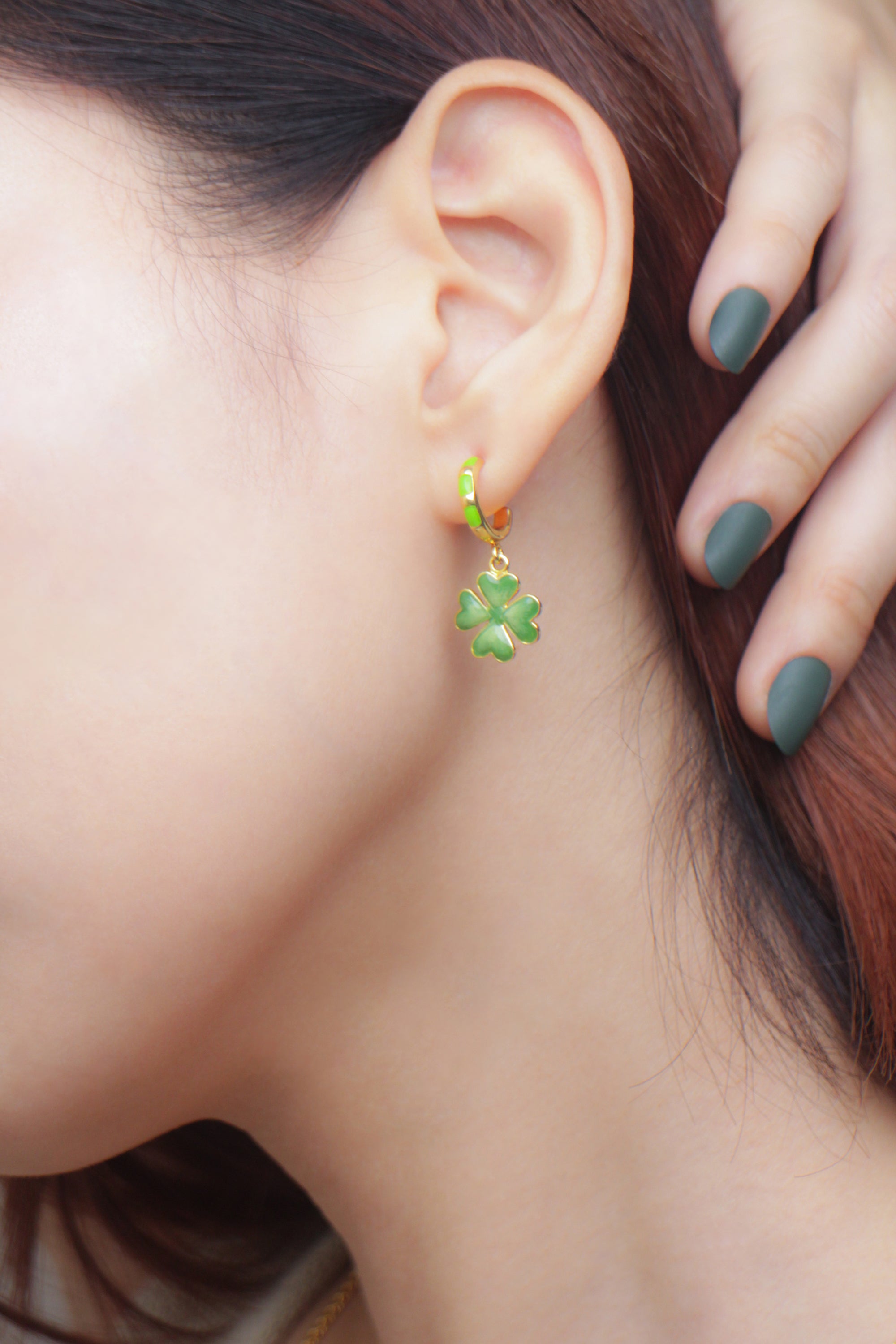 A close-up of a woman's ear showcasing a green clover earring and her manicured dark green nails.