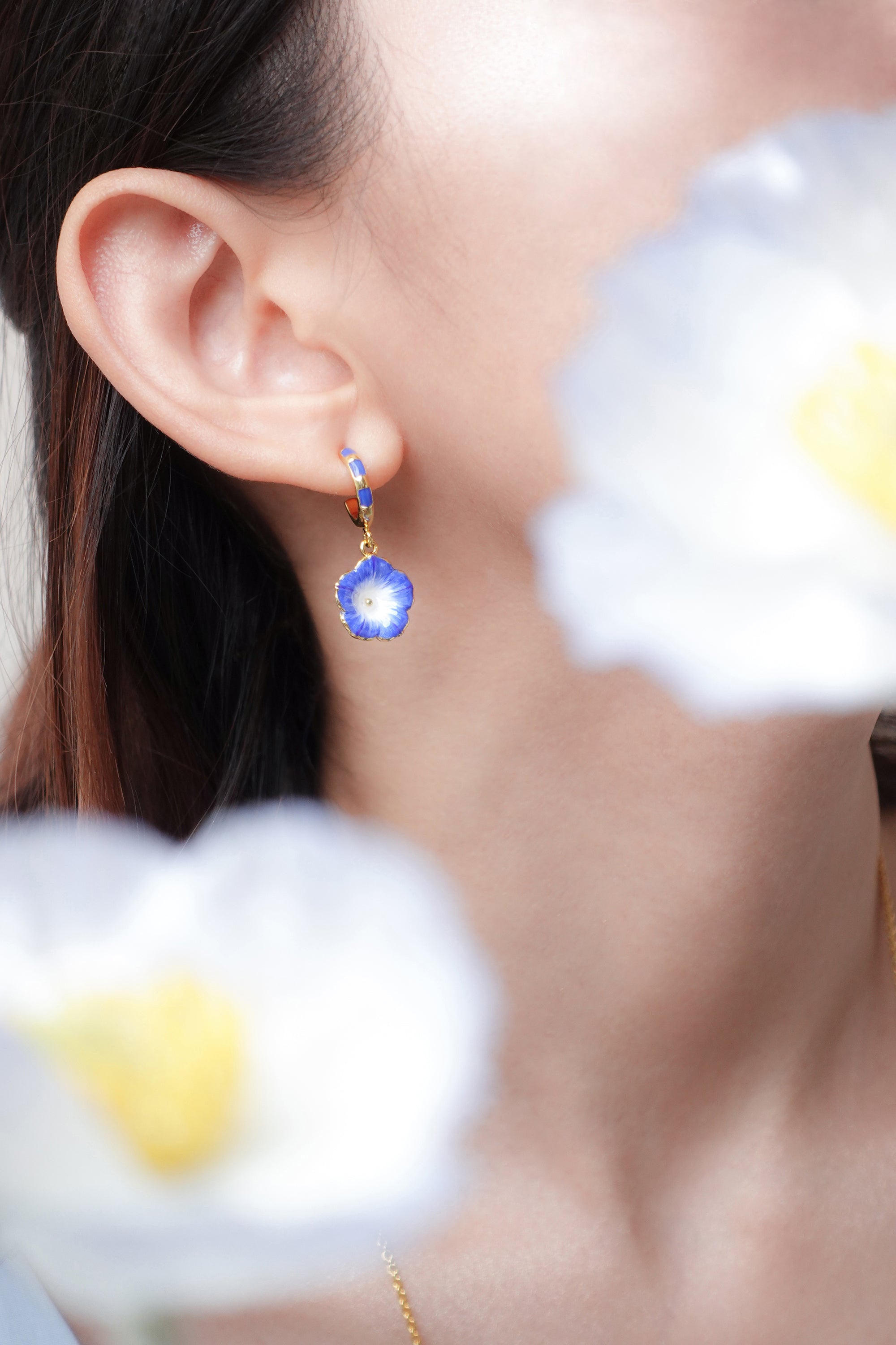 A close-up of a woman wearing blue floral earrings, partially obscured by white flowers.