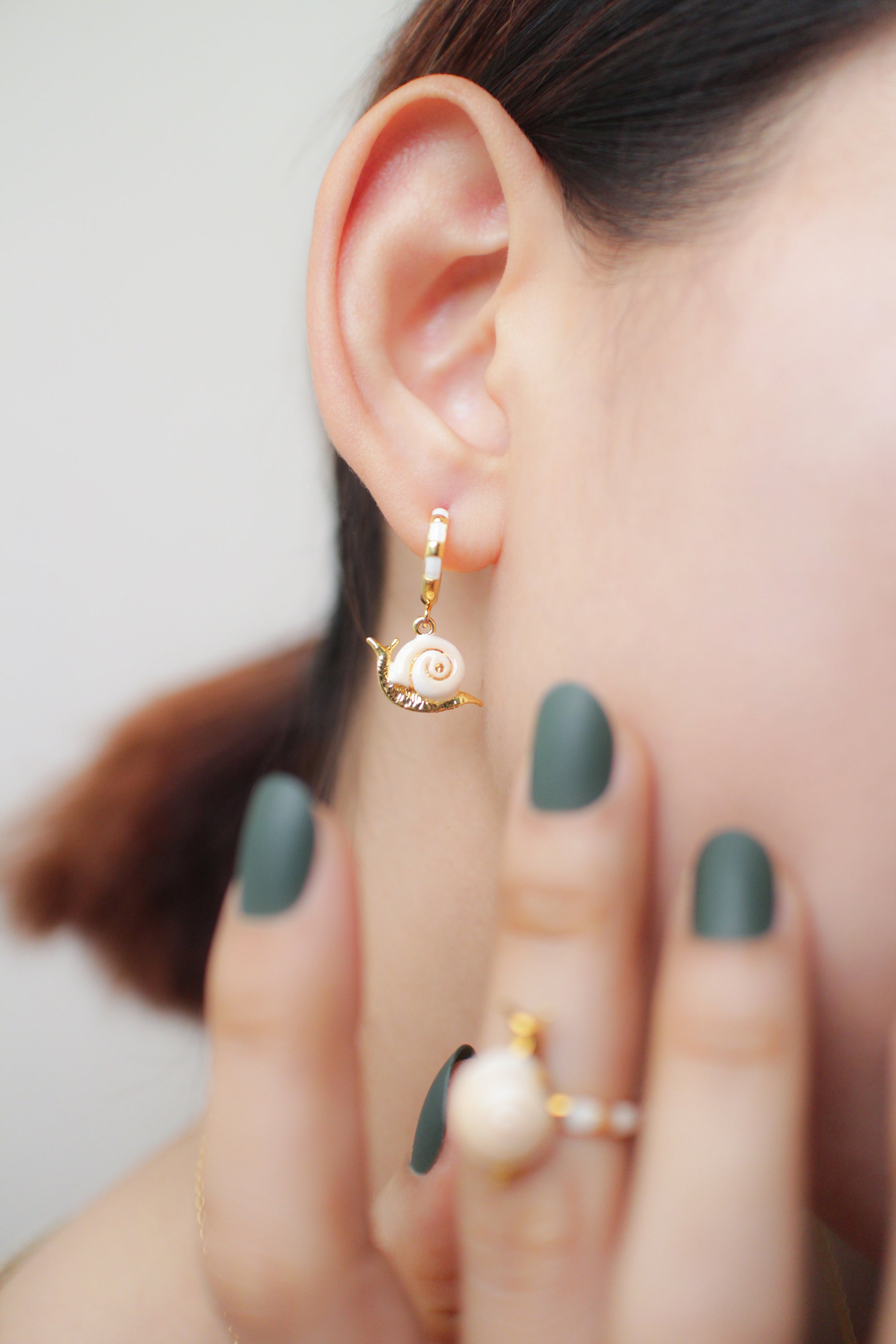 Gold snail earrings worn by a person with green nails, partially obscured by their hand.