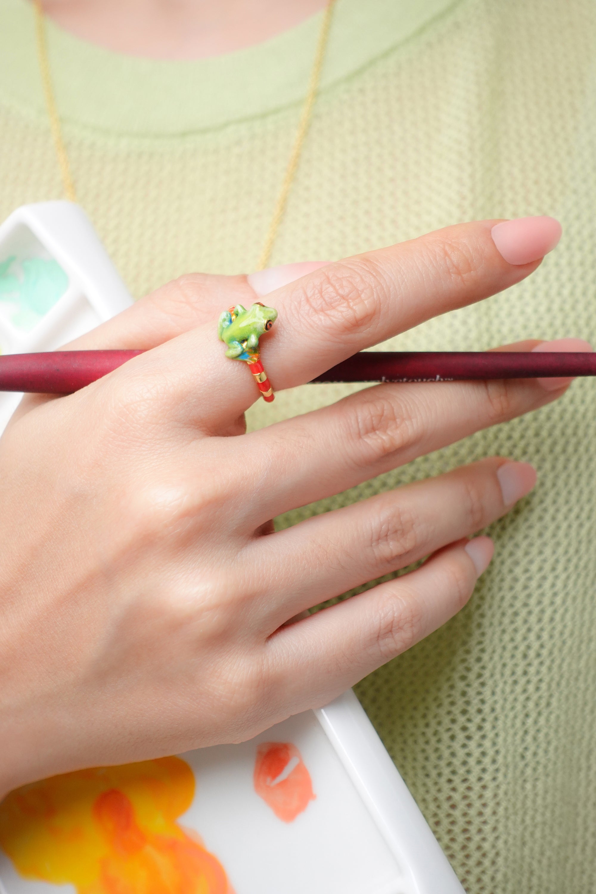 A colorful frog ring worn on a person's hand holding a paintbrush.