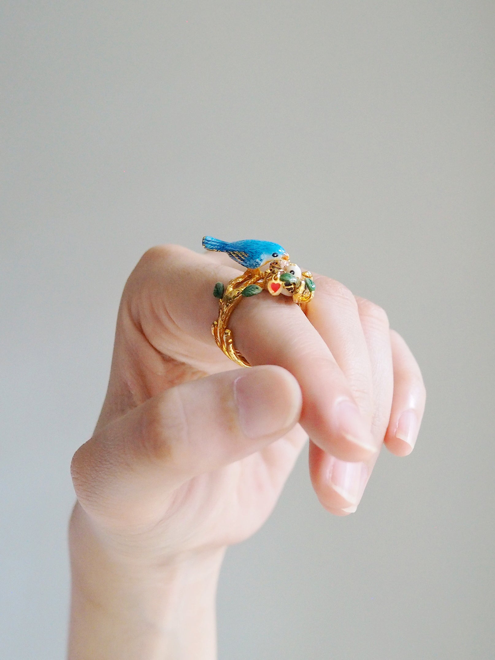 Close up of a hand showcasing a gold ring featuring blue bird and a baby bird in the nest.