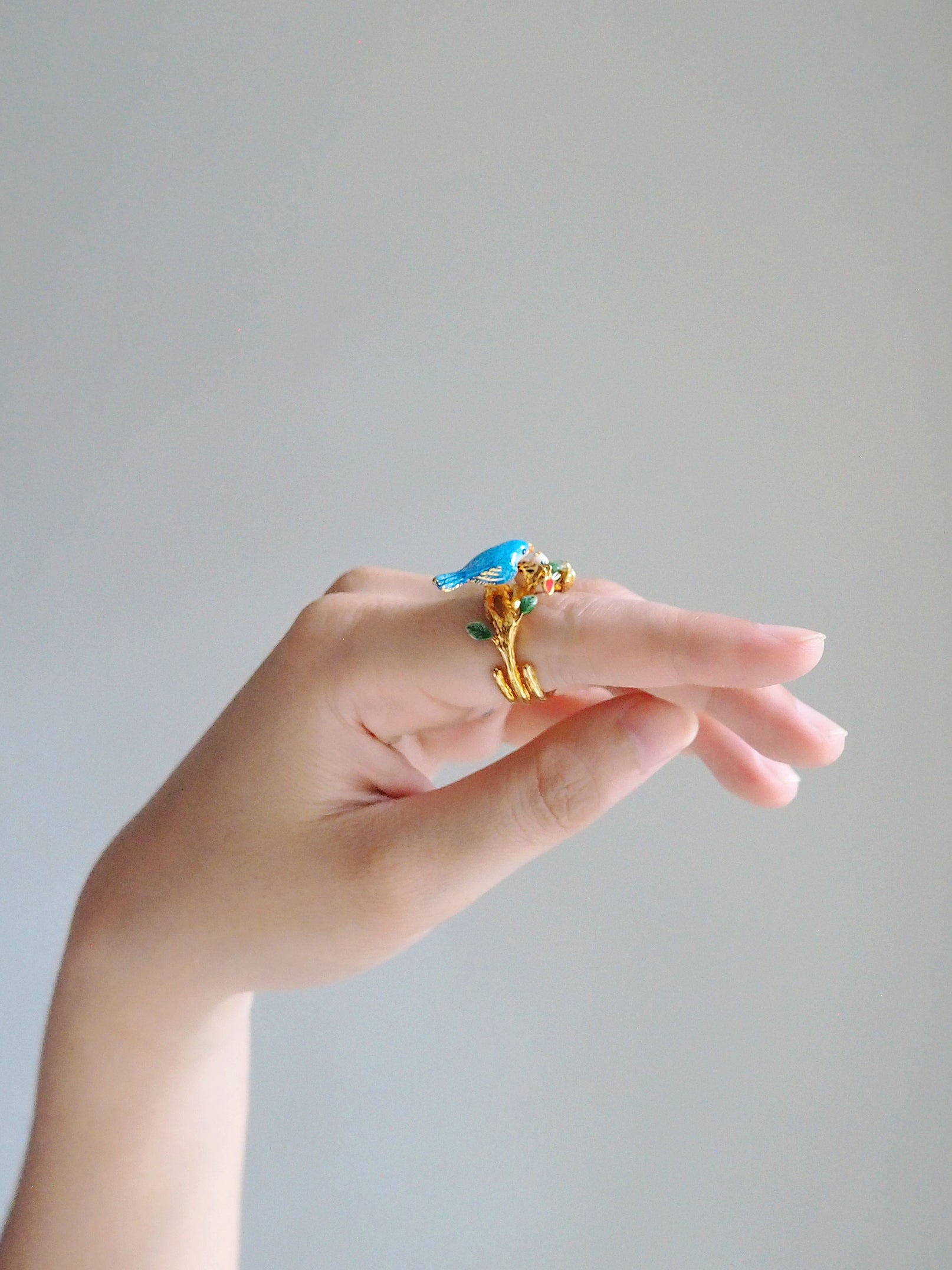 Close up of a hand showcasing a gold ring featuring blue bird and a baby bird in the nest.