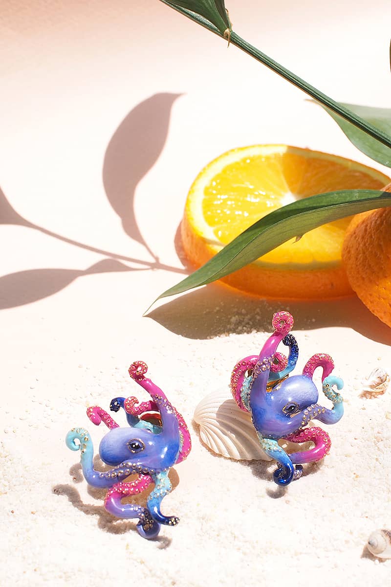 Colorful octopus earrings placed on sandy surface, with orange slices and a shell in the background.