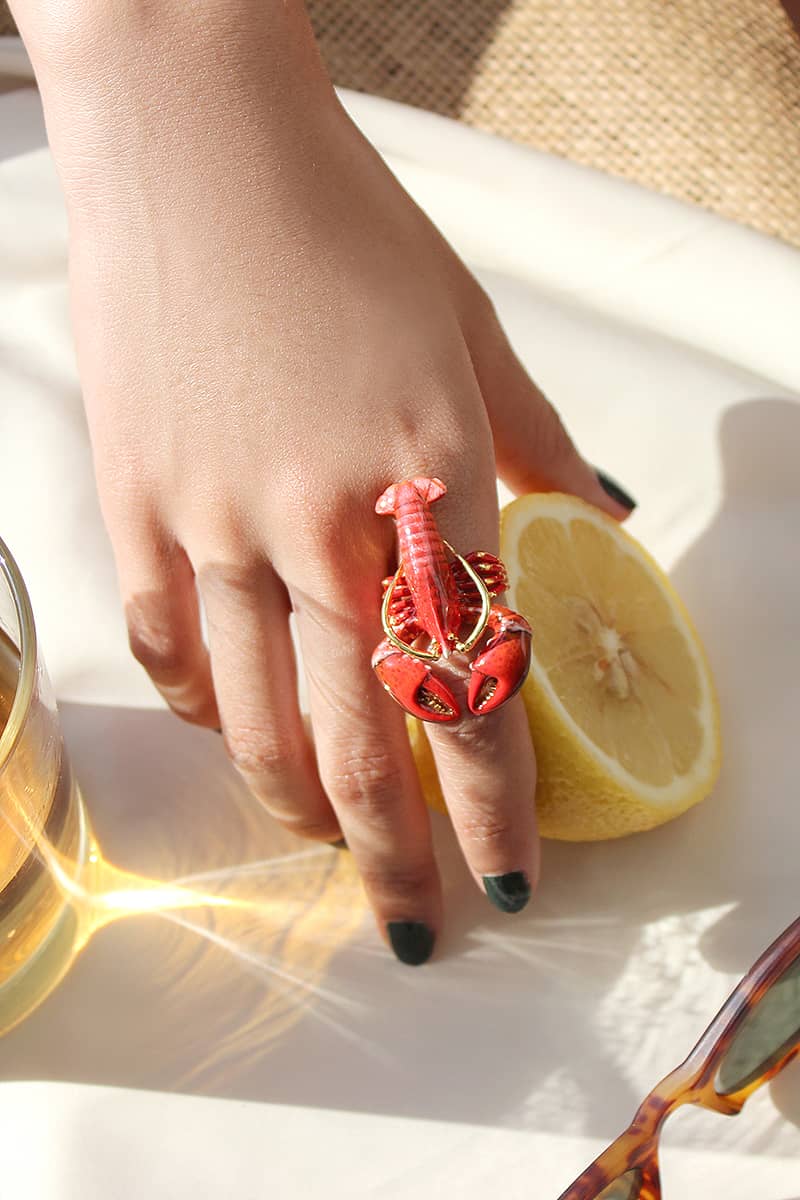 A close-up of a red lobster ring worn on a hand, while grabbing a lemon.