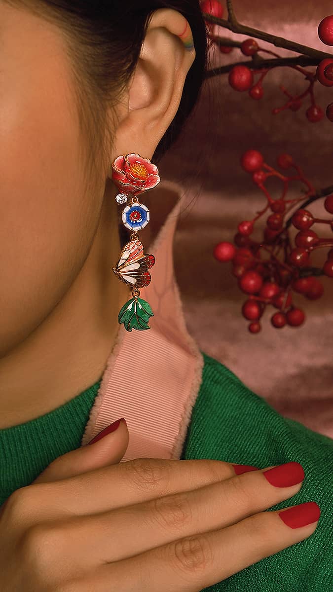 Colorful flower-shaped and butterfly earrings worn by a person with red nails, holding a pink strap.