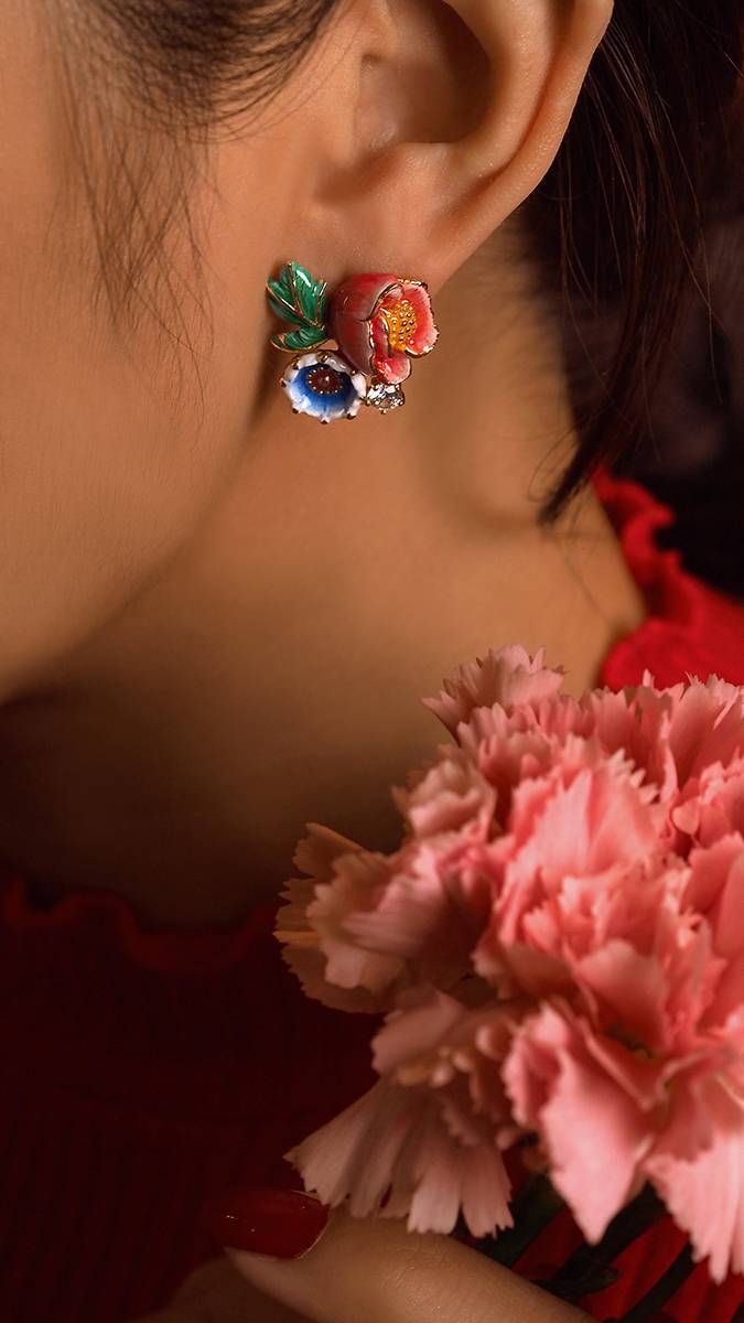 A close-up of a woman's ear adorned with colorful floral earrings, complemented by a pink carnation nearby.