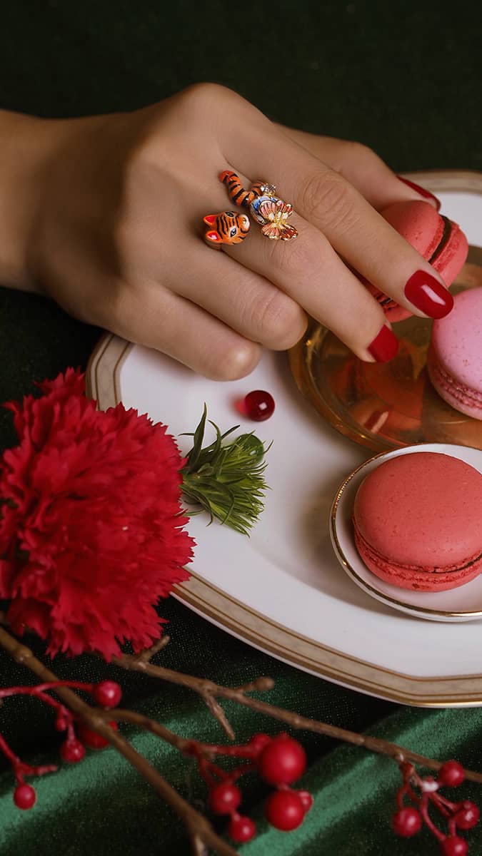 A hand with a tiger ring with a butterfly and floral elements, holds a pink macaron, surrounded by flowers and berries.